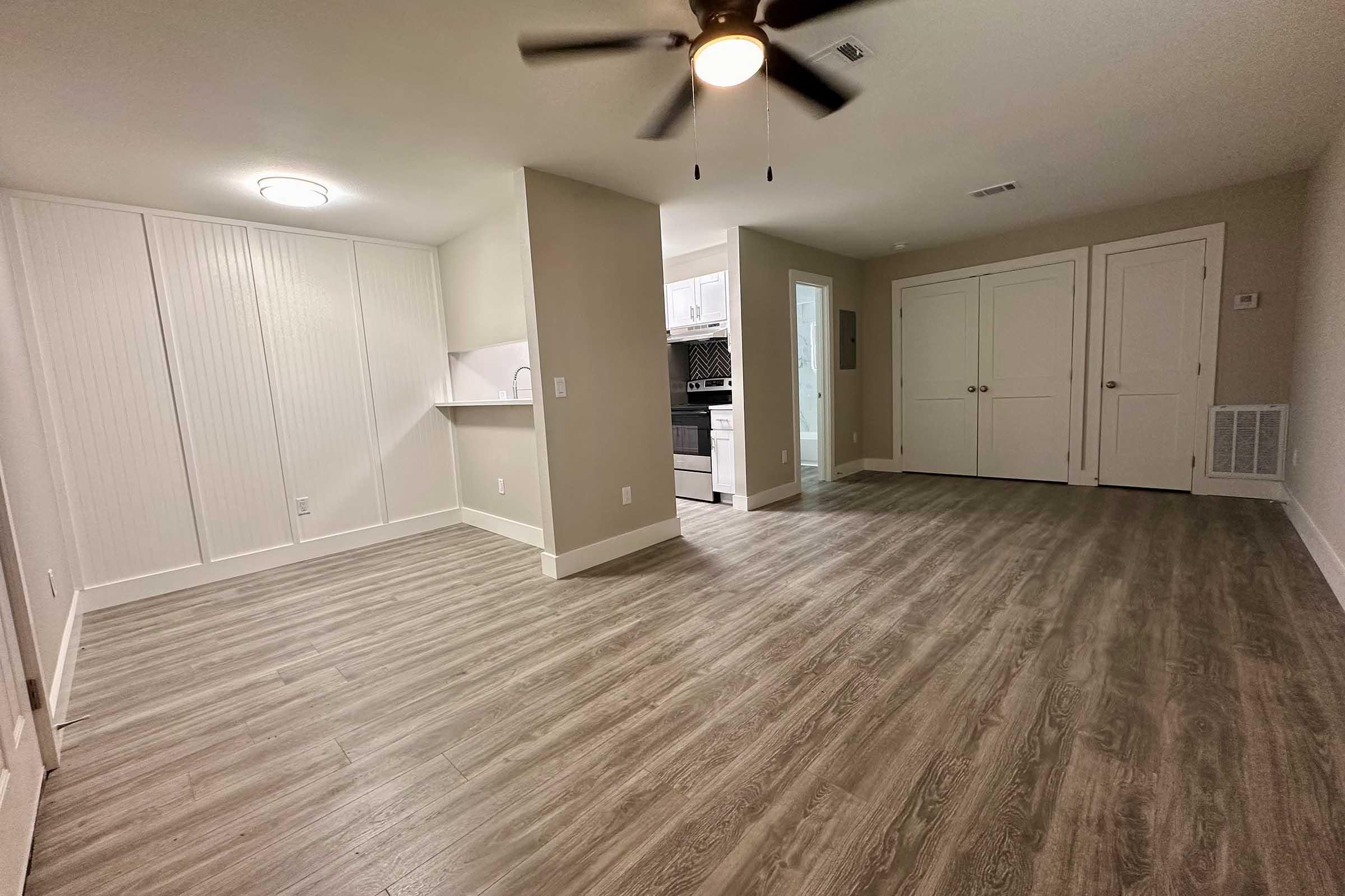 Spacious interior of an apartment featuring hardwood-style flooring, a ceiling fan, and a neutral color palette. The room includes a kitchenette area visible in the background, along with two closed doors on the right. Ample natural light brightens the space.