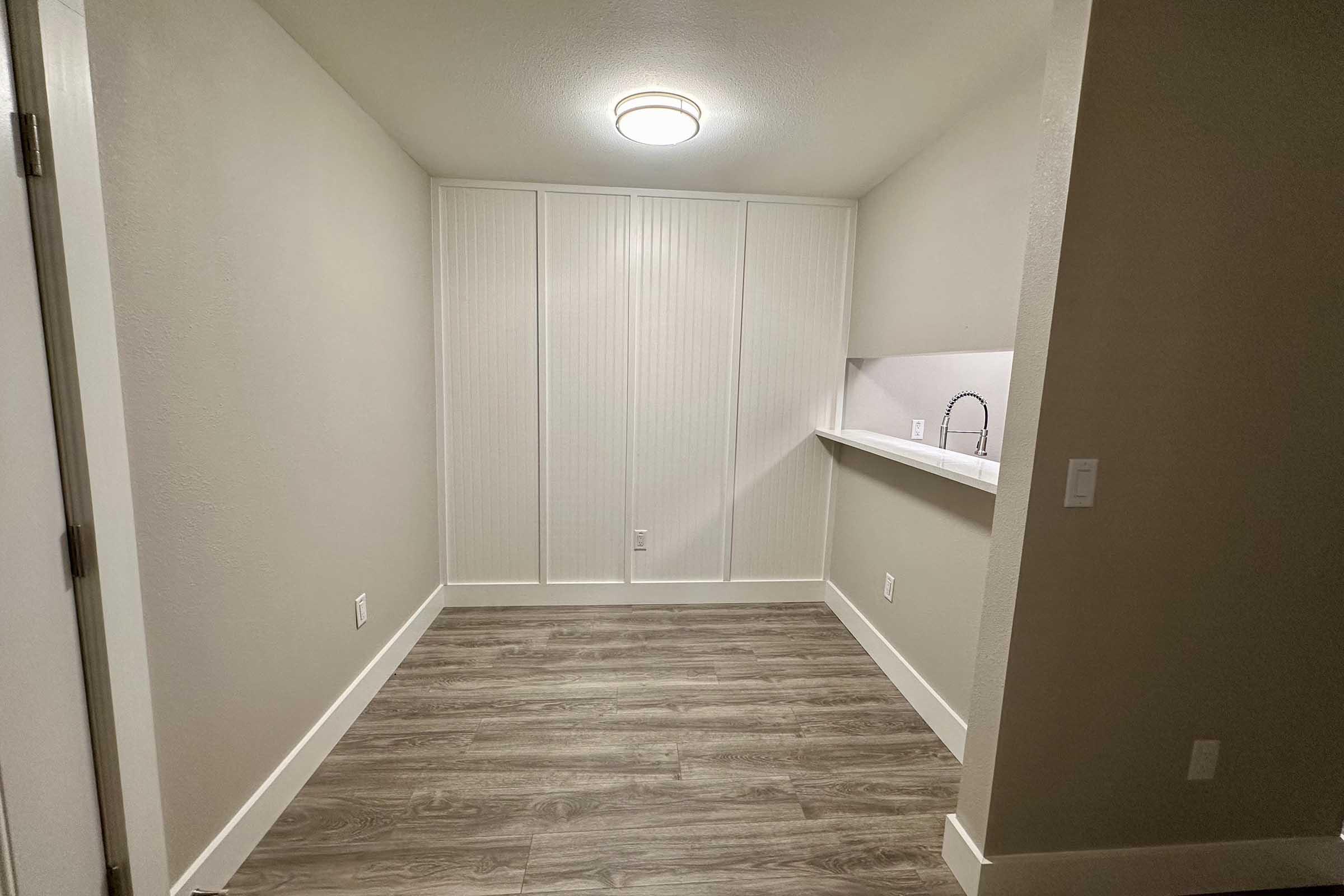 A small, empty room with light gray walls and wood-like flooring. There is a smooth countertop and a wall with a subtle vertical panel design. The ceiling has a modern light fixture, and the space is well-lit, providing a clean and minimalistic aesthetic.
