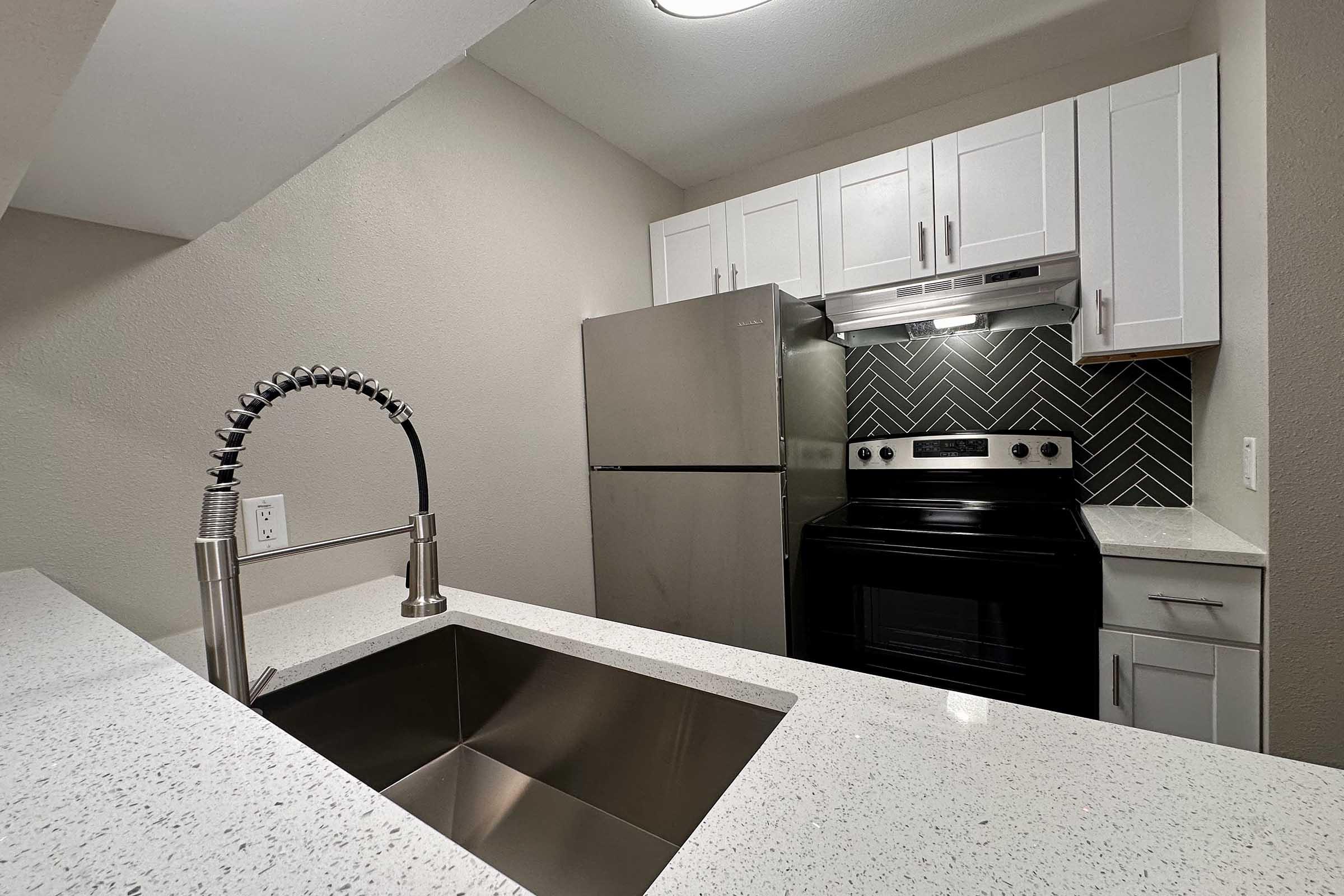 A modern kitchen featuring white cabinets, a stainless steel refrigerator, and an oven with a black range. The countertop is light-colored with a sleek design, and there is a stylish sink with a pull-down faucet. The backsplash is patterned with herringbone tiles, creating a contemporary look.