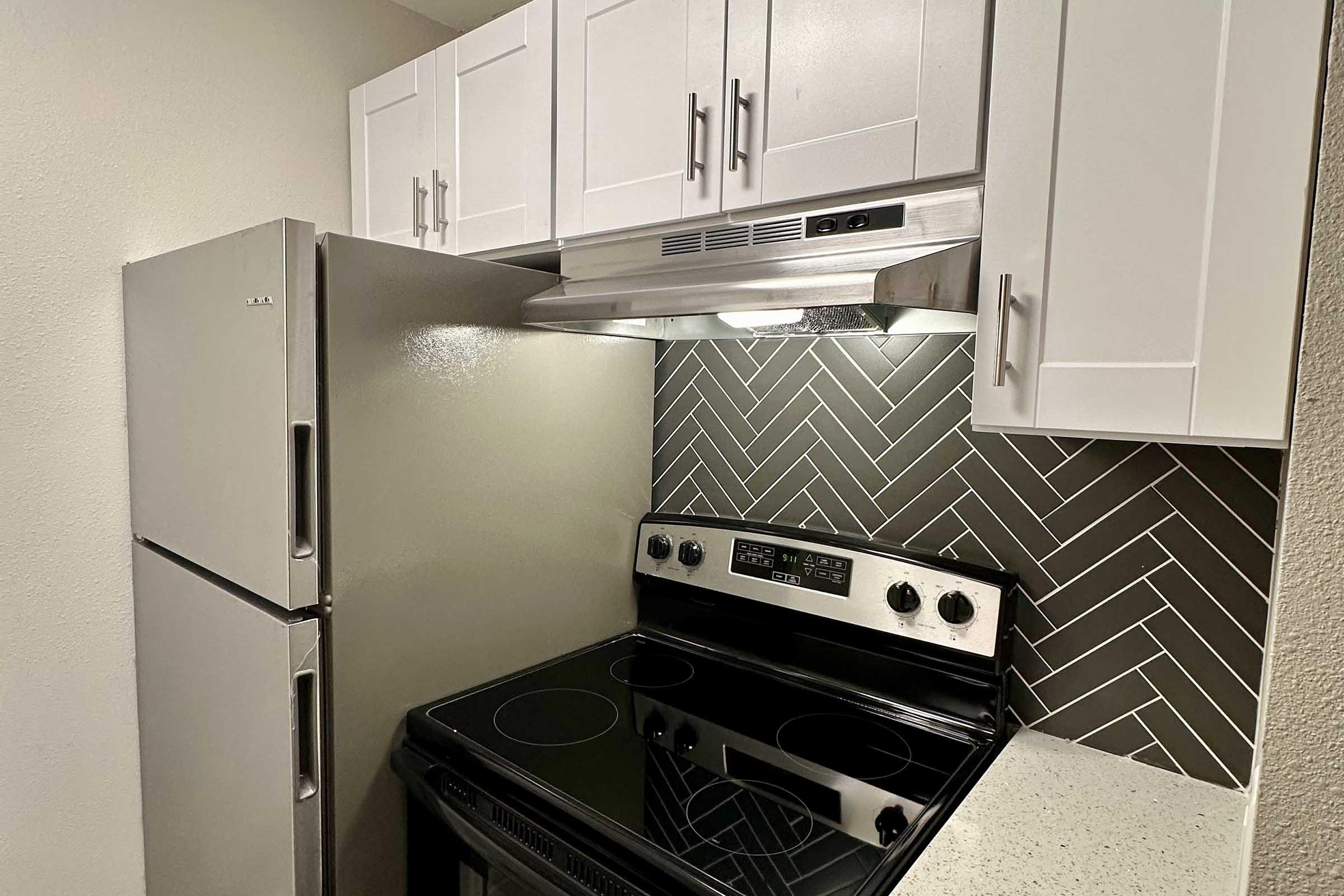 A modern kitchen featuring a stainless steel refrigerator, black electric stove with an oven, and a white tiled backsplash arranged in a herringbone pattern. The cabinets above are white, complementing the sleek design of the kitchen space. The countertop appears light and textured.