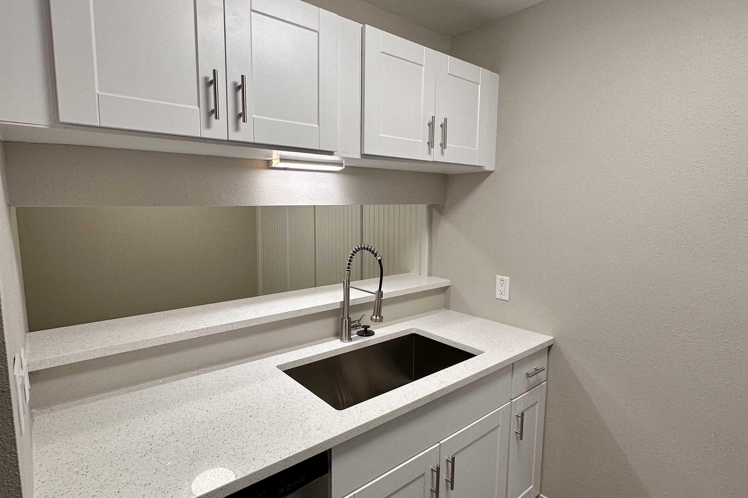 A modern kitchen space featuring white cabinetry, a silver faucet above a stainless steel sink, and a smooth, light-colored countertop. The walls are painted in a neutral tone, providing a clean and minimalistic aesthetic.