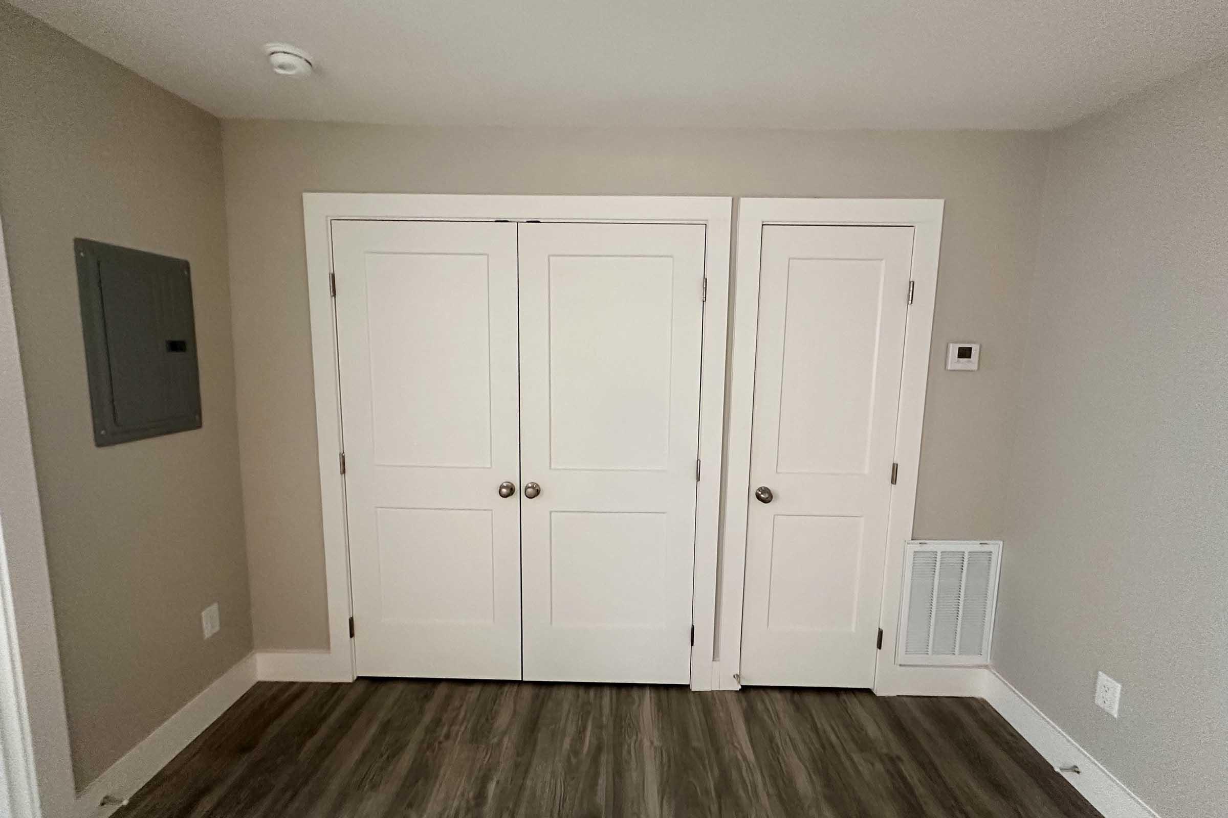 A small room featuring two sets of white double doors, with one door possibly leading to a closet. The walls are painted a light gray, and the flooring is a light wood-like material. There’s a wall-mounted electrical panel on the left and a thermostat on the right.