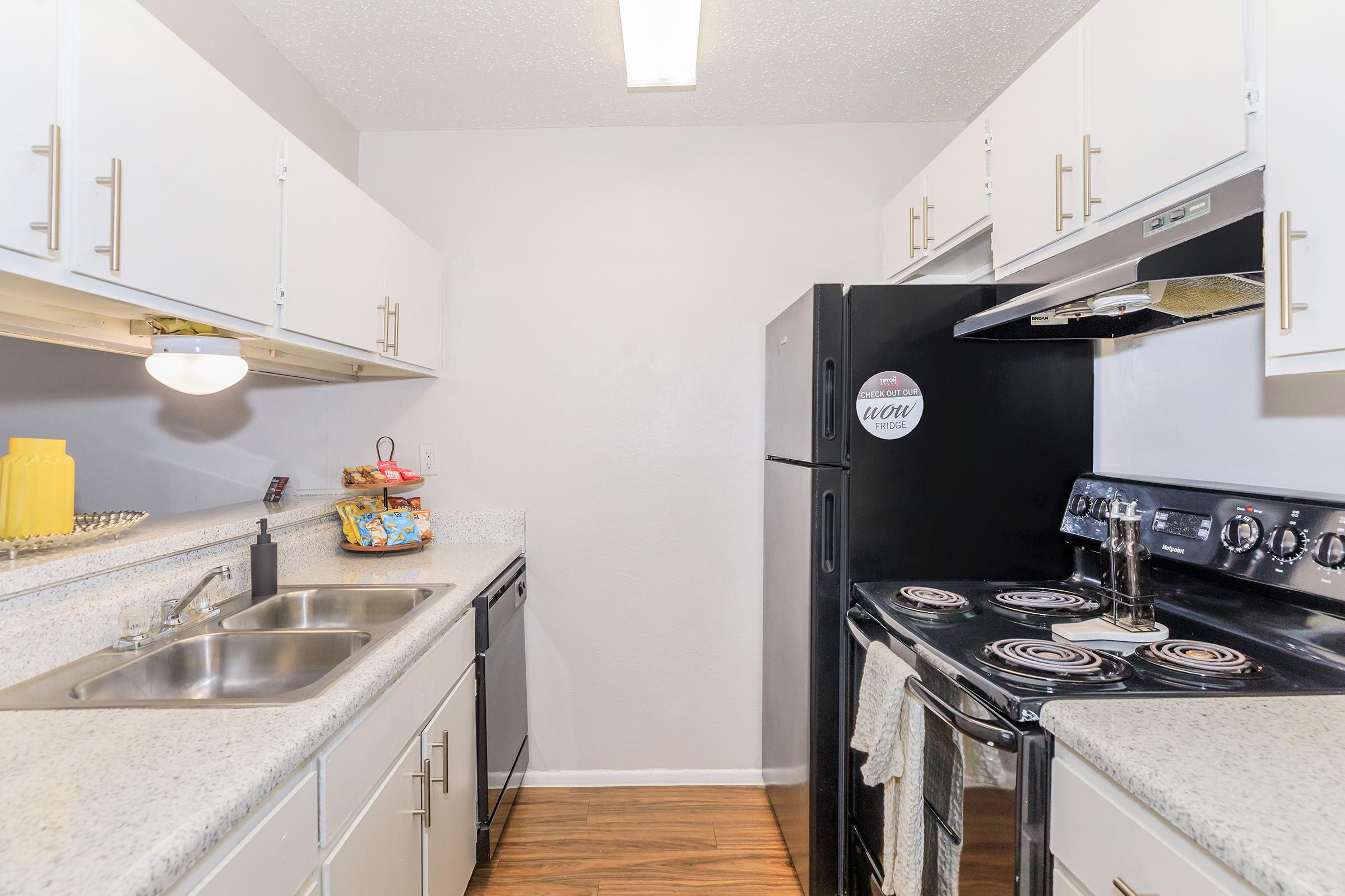 A modern kitchen featuring white cabinets, a black refrigerator, and a black stove with an oven. The countertops are light granite, and there is a double sink beneath a bright light fixture. The kitchen is well-organized, with snacks displayed on the counter and towels hanging on the stove.