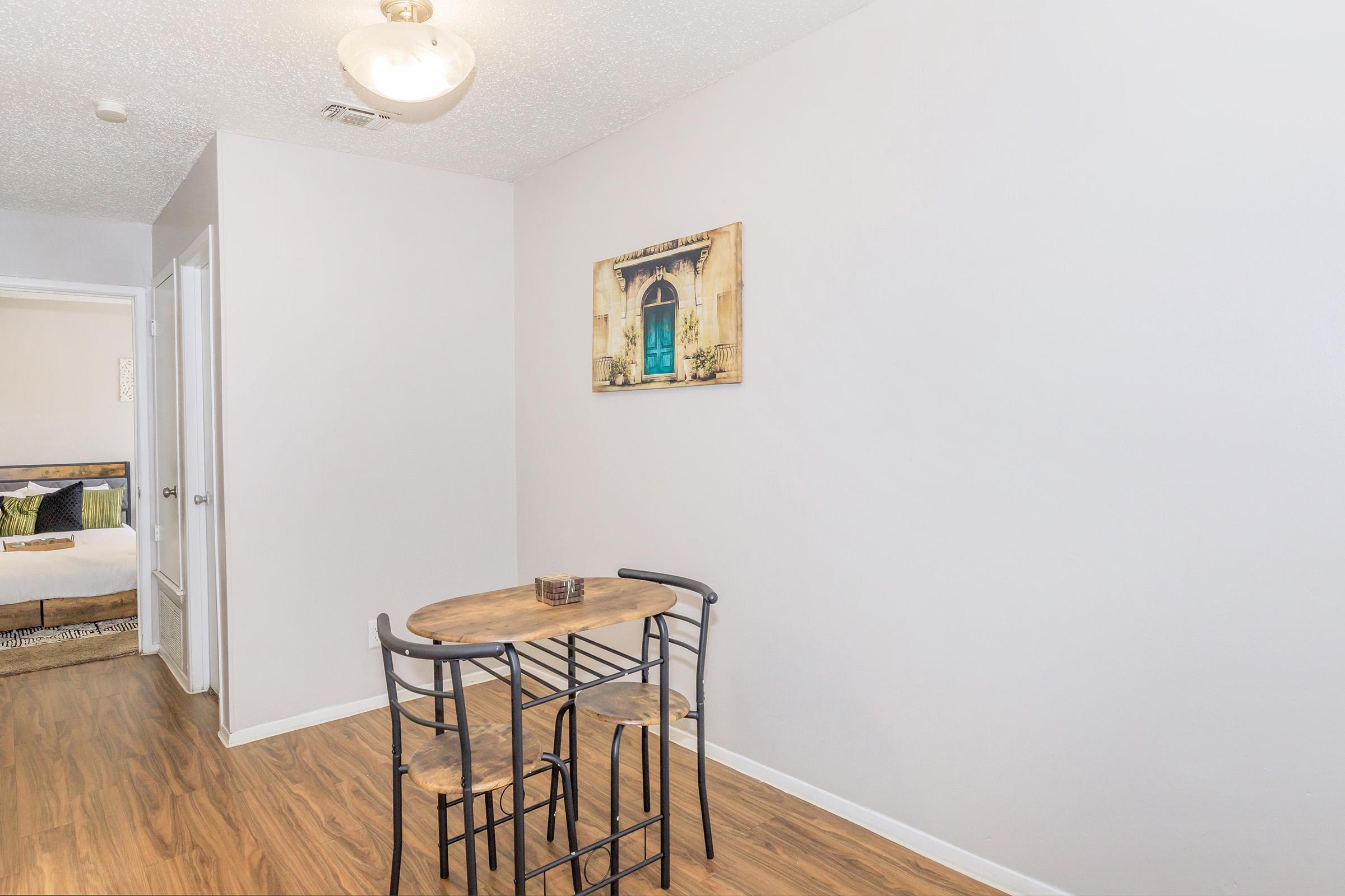 A small dining area featuring a round wooden table with black metal chairs. A decorative item sits on the table. The walls are painted light gray, and a piece of art depicting a window with green shutters hangs on one wall. The flooring is a warm wood finish.
