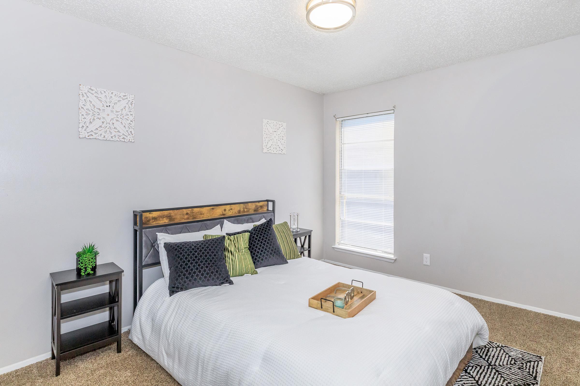A neatly arranged bedroom featuring a queen-sized bed with white bedding and decorative pillows. There is a small bedside table with a plant, and the walls are adorned with simple artwork. A window allows natural light to enter, and the carpeted floor adds a warm touch to the space.