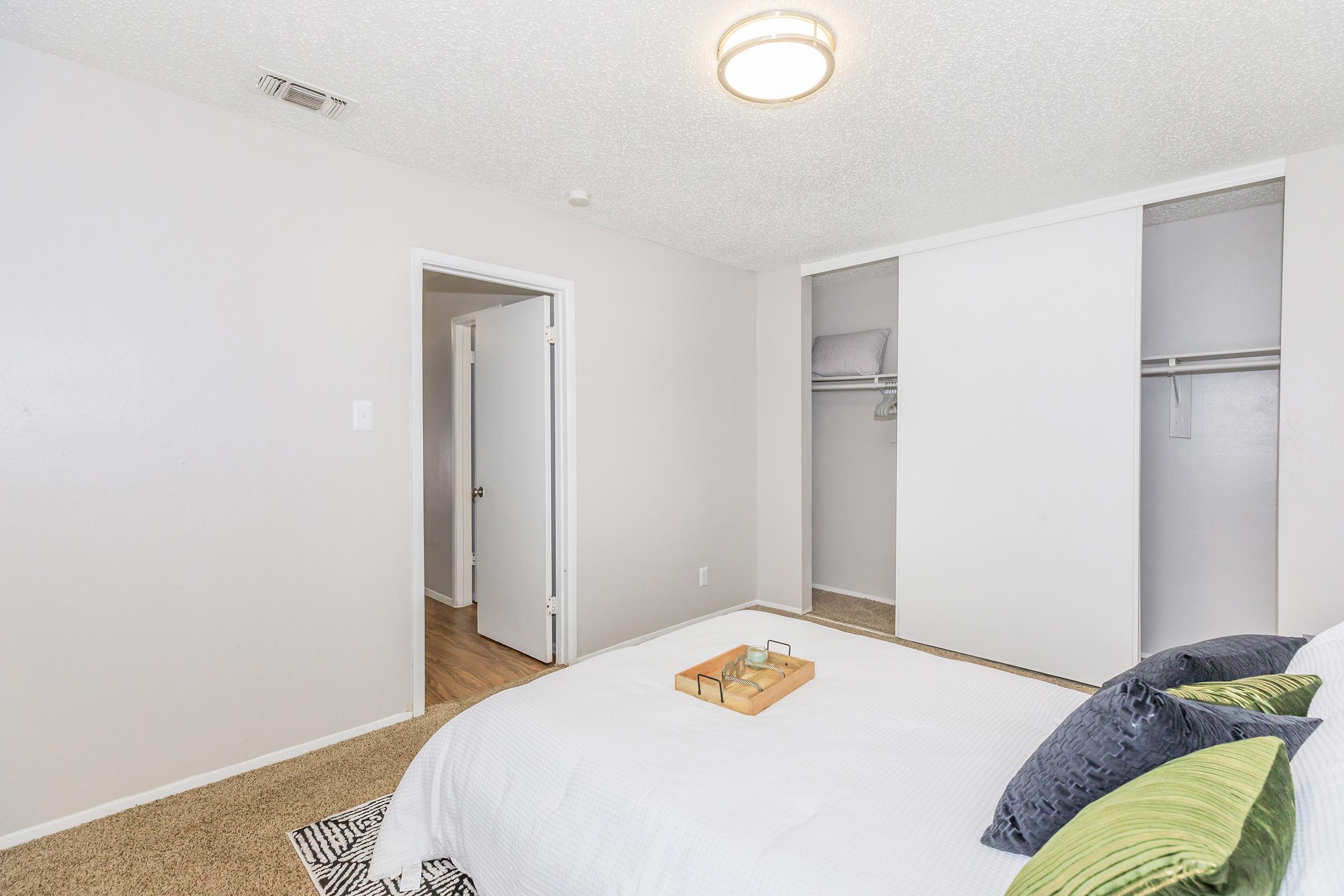 A neatly arranged bedroom with a bed featuring decorative pillows, a tray on top, and light-colored walls. A closet with sliding doors is visible, and a door leads to another room. The floor is wooden, adding a modern touch to the space. The lighting is soft and warm.