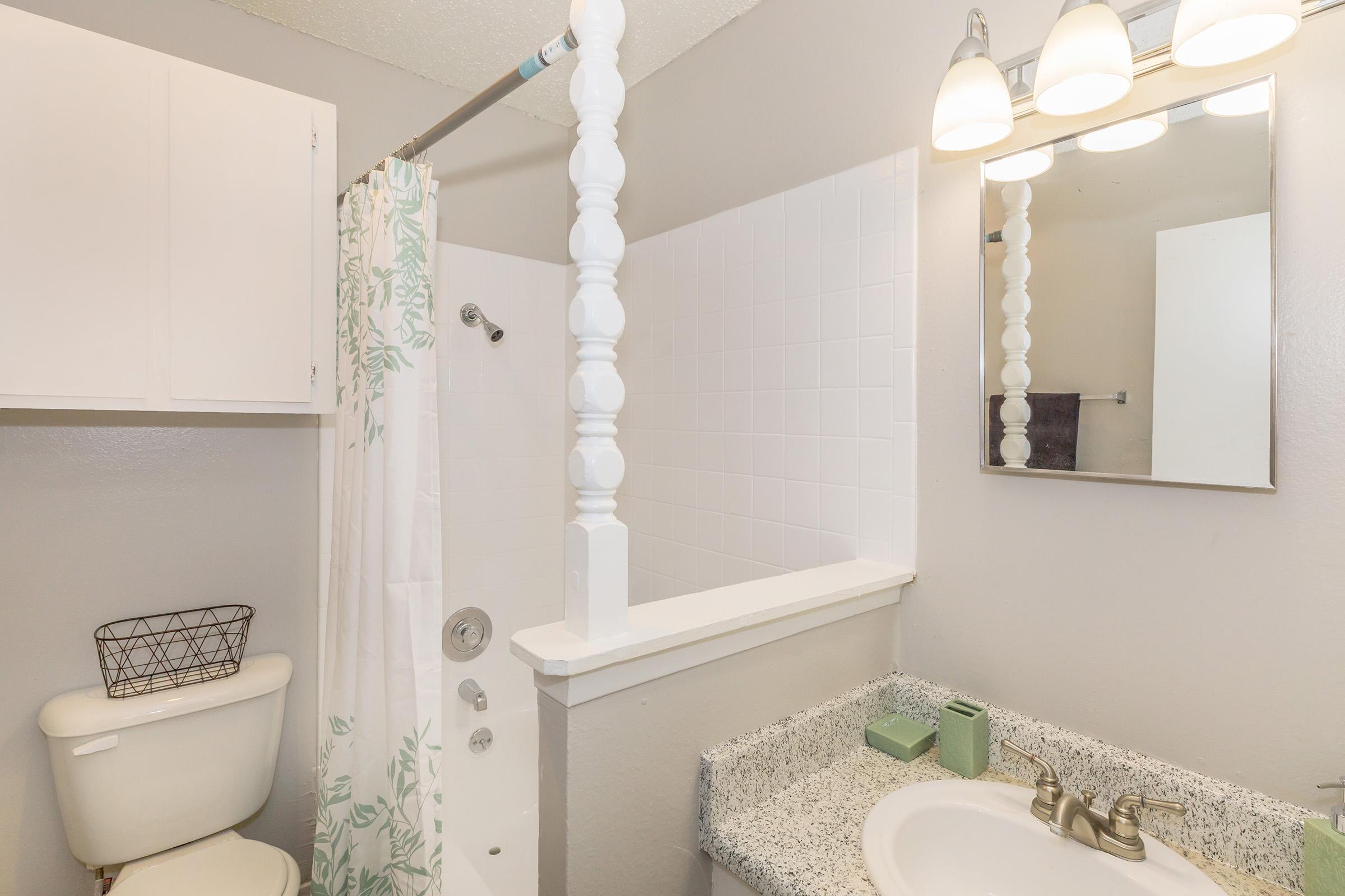 A bathroom featuring a toilet, a shower area with a curtain, a sink with a speckled countertop, and a mirror. The walls are painted light gray, and there are overhead lights illuminating the space. A small wall-mounted cabinet is shown above the toilet.