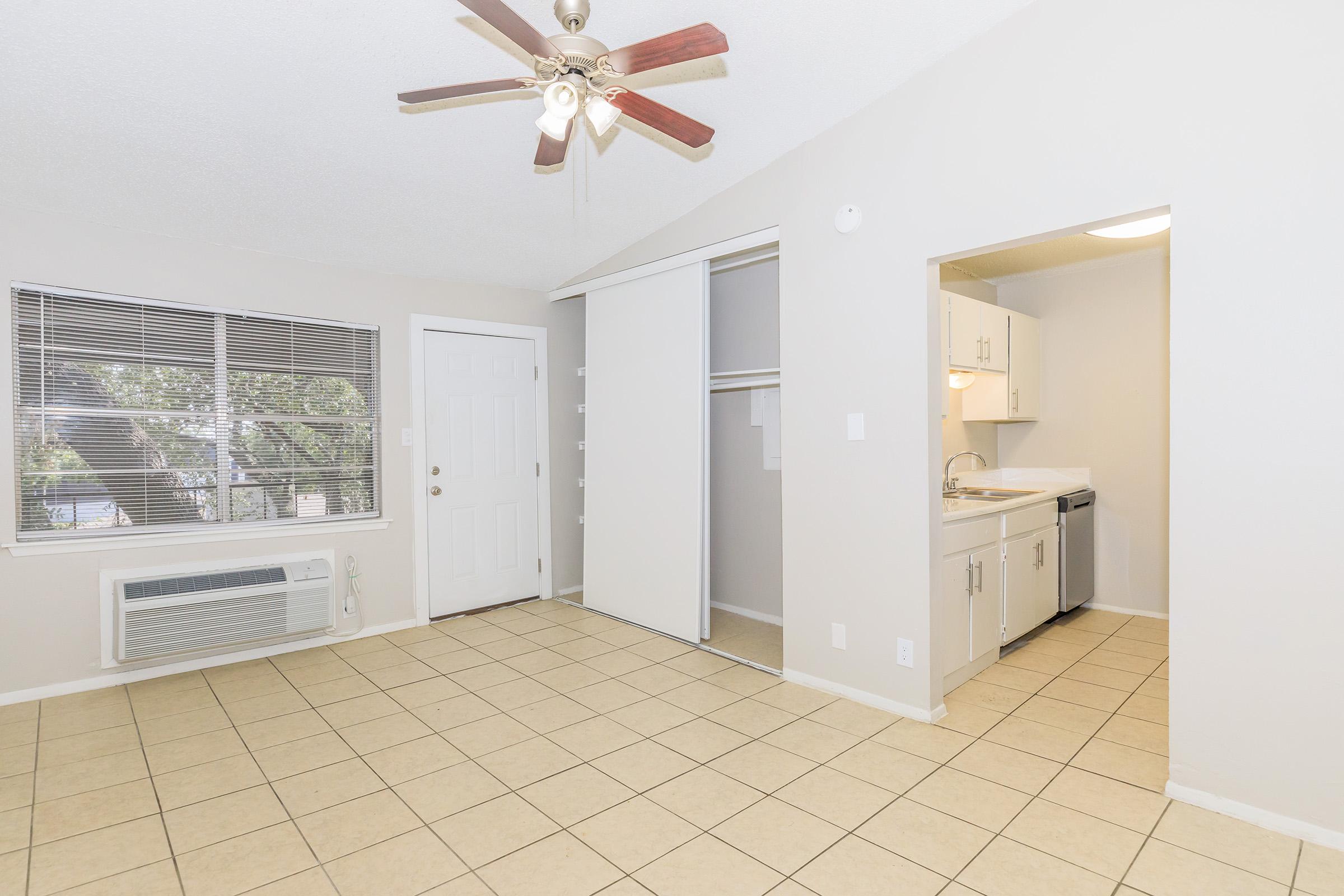 A small, brightly lit living space featuring beige tile flooring, a ceiling fan, and an air conditioning unit. There is a sliding door closet on one side and a kitchenette with white cabinets and a sink in the background. Large windows provide natural light, and the walls are painted light grey.