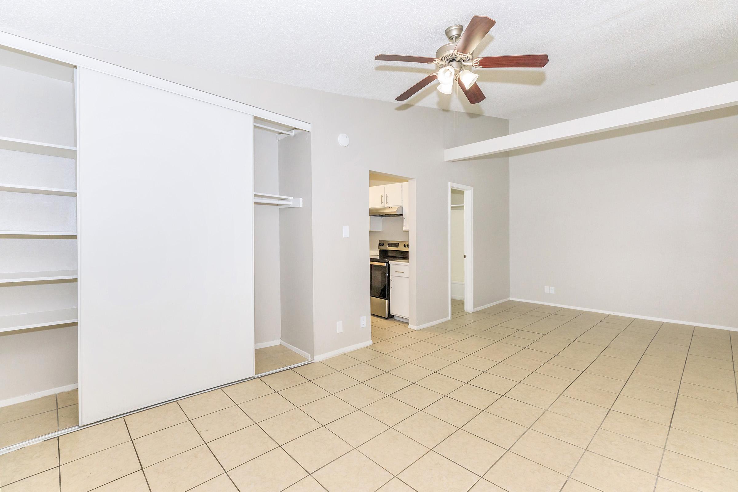 Interior view of a spacious apartment living area featuring a ceiling fan, beige walls, and tiled flooring. A closed closet is visible to the left, and there is an open kitchen area in the background. The layout appears bright and inviting, suitable for comfortable living.