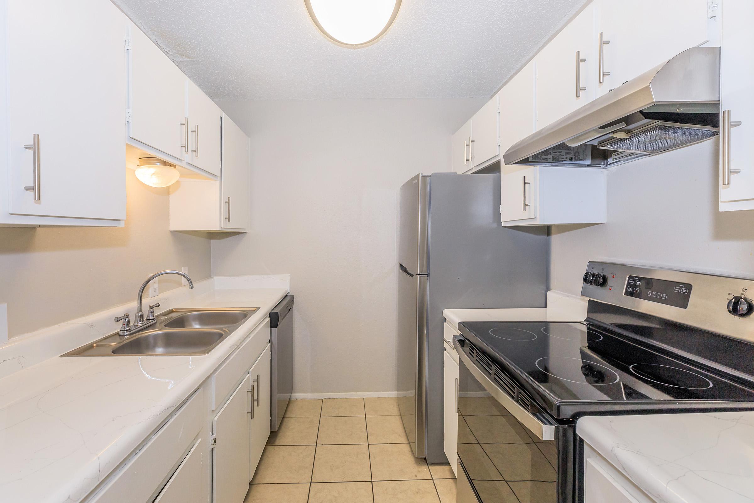 A modern, well-lit kitchen featuring white cabinetry, a double sink, a stainless steel refrigerator, and a black electric stove with an overhead vent. The countertops are light-colored and the flooring is tiled. The walls are painted in a soft neutral color, enhancing the space's brightness.