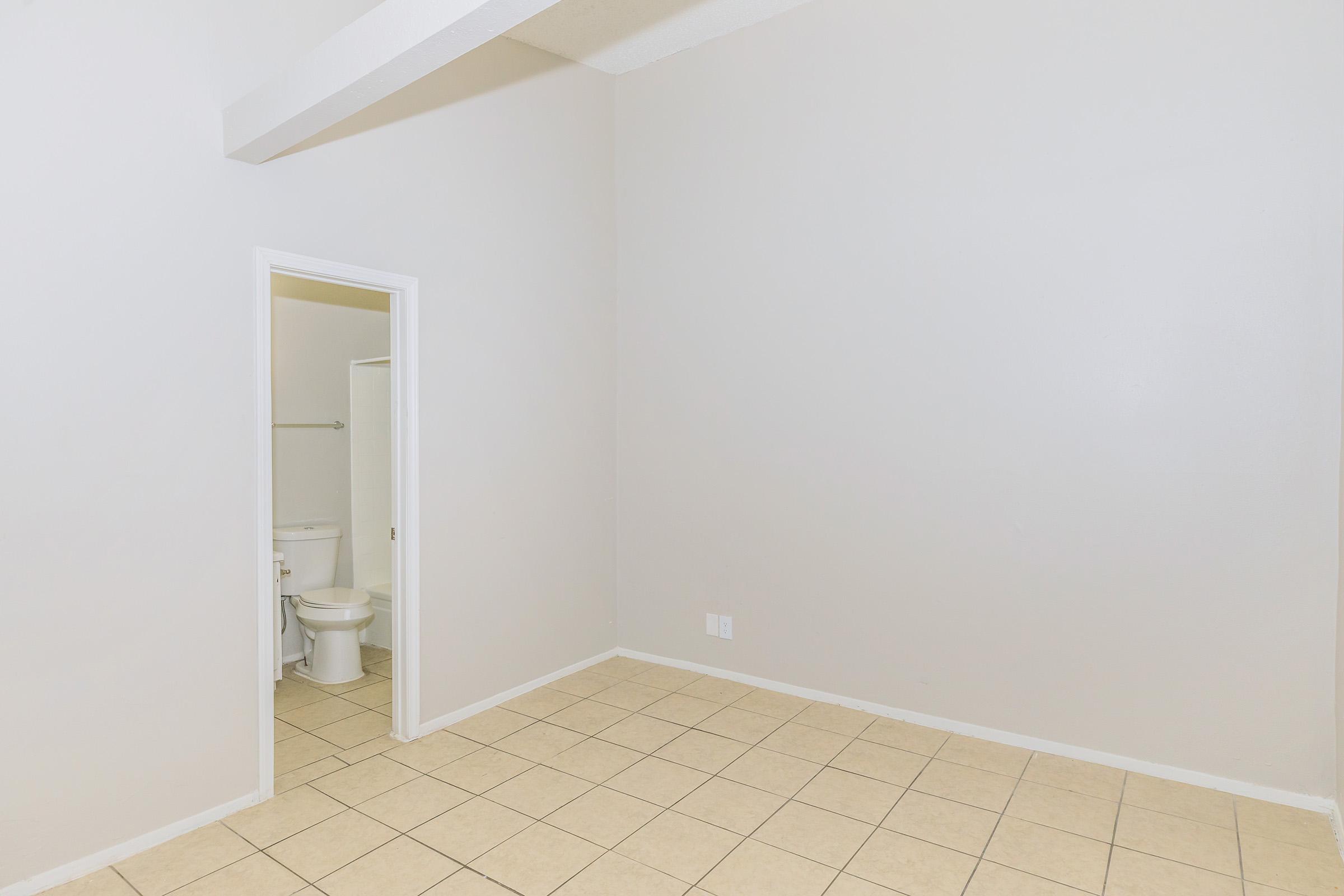 An empty room with light gray walls and tiled flooring. There is a doorway leading to a bathroom visible in the corner, featuring a toilet. The ceiling has exposed beams, and the room is well-lit, creating a spacious feel.