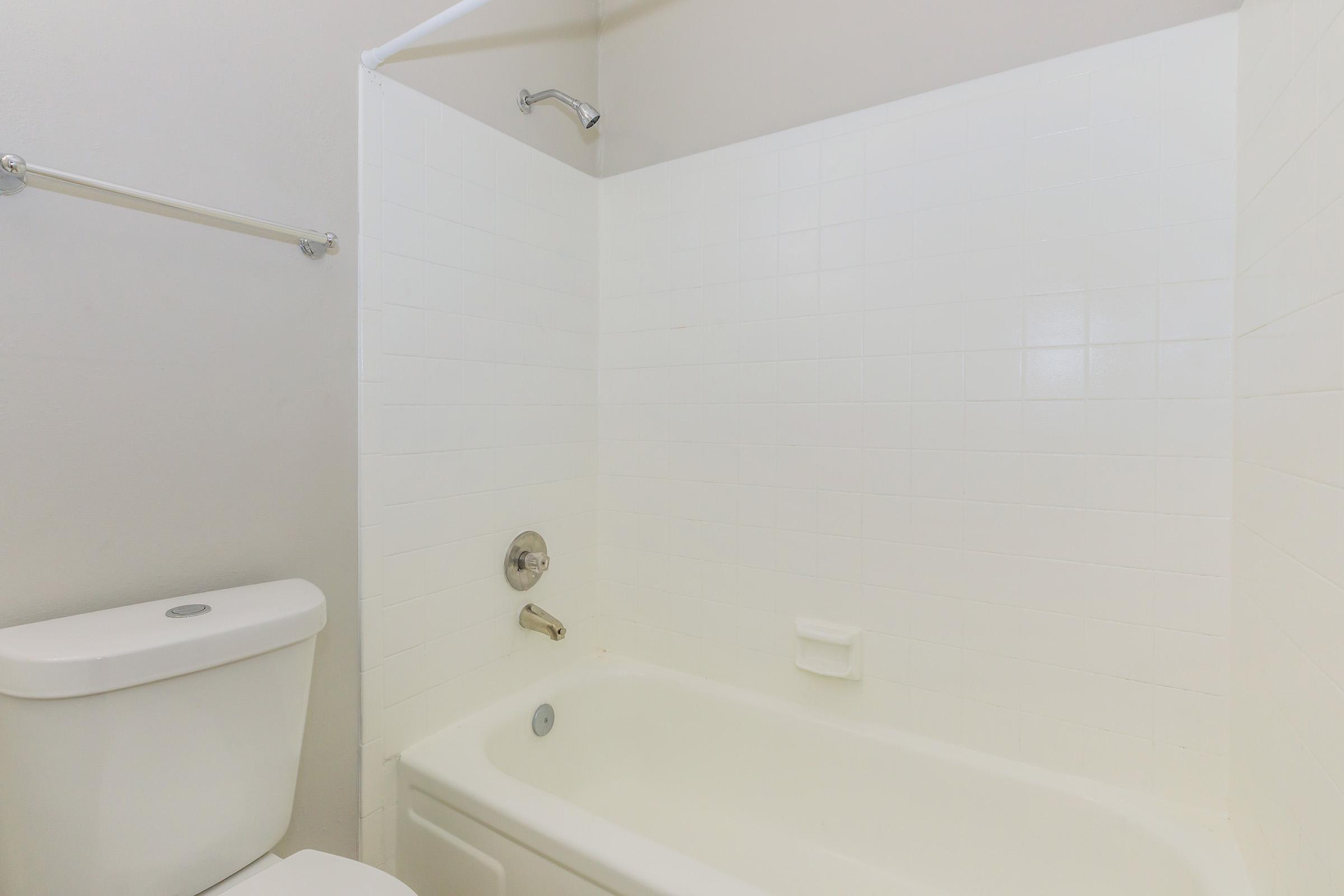 A clean, simple bathroom featuring a white bathtub, a showerhead, a wall-mounted towel rack, and a toilet. The tiled walls are light-colored, creating a bright and airy atmosphere. The overall design is minimalist with a focus on functionality.