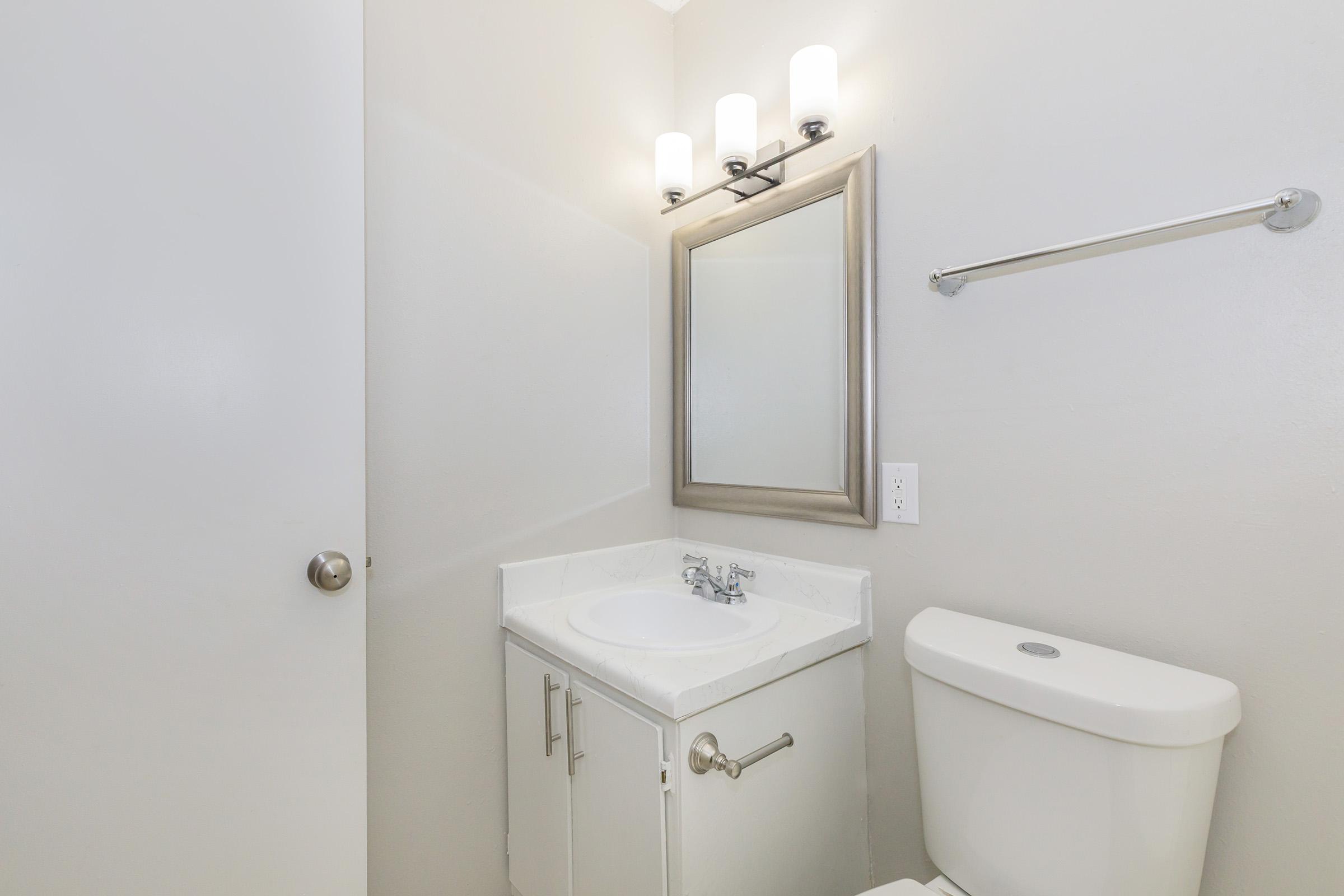 A small bathroom featuring a white sink with a mirror above, a toilet on the right, and wall-mounted lights. The walls are painted a soft gray, and there is a towel rack. A door is partially visible on the left. The overall space is clean and modern.