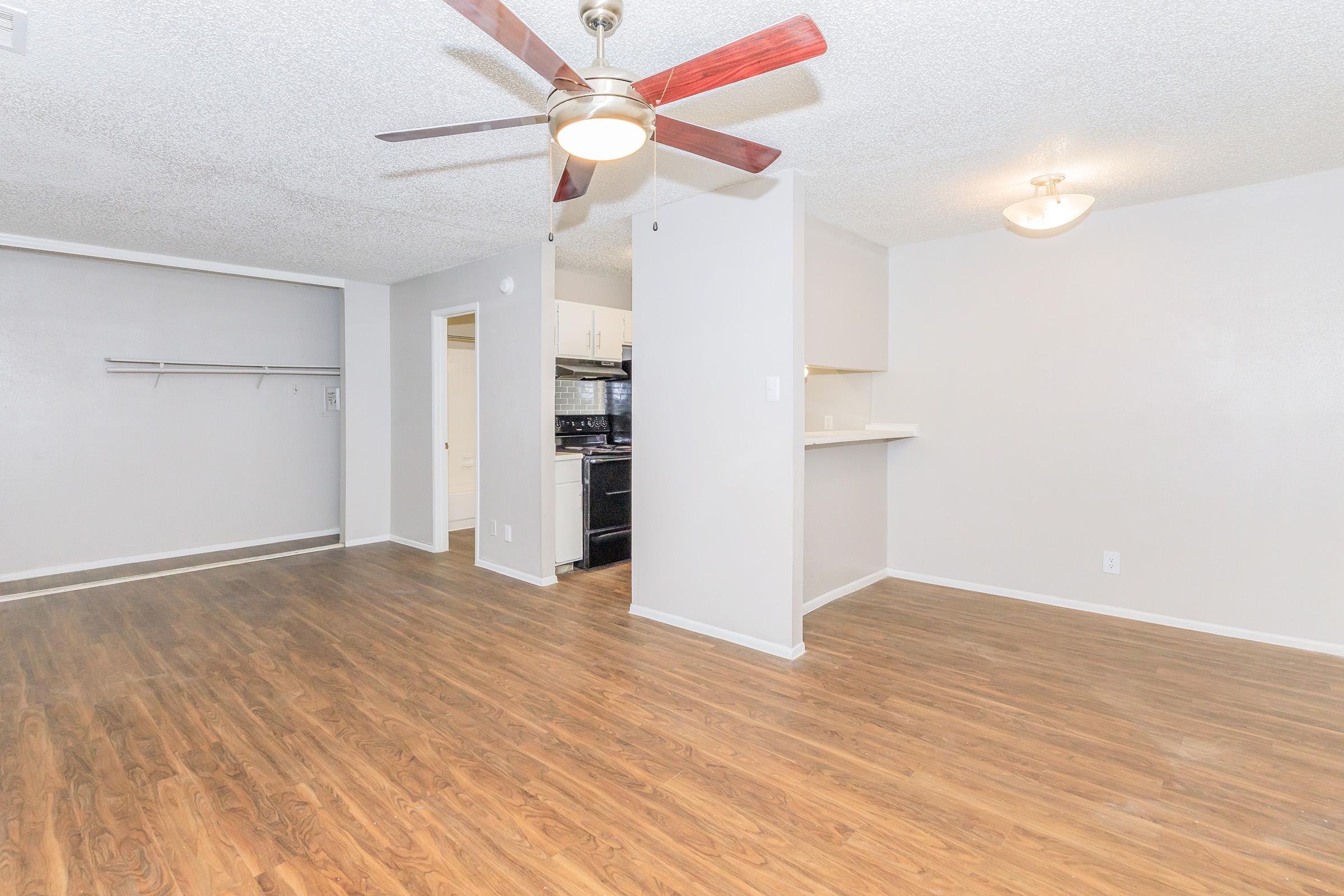 A spacious, well-lit living area featuring wood-style flooring, a ceiling fan, and light fixtures. The room has neutral gray walls and a small bar area leading into a kitchen, which is not fully visible. There's a coat rack on the left, enhancing the open layout of the space.