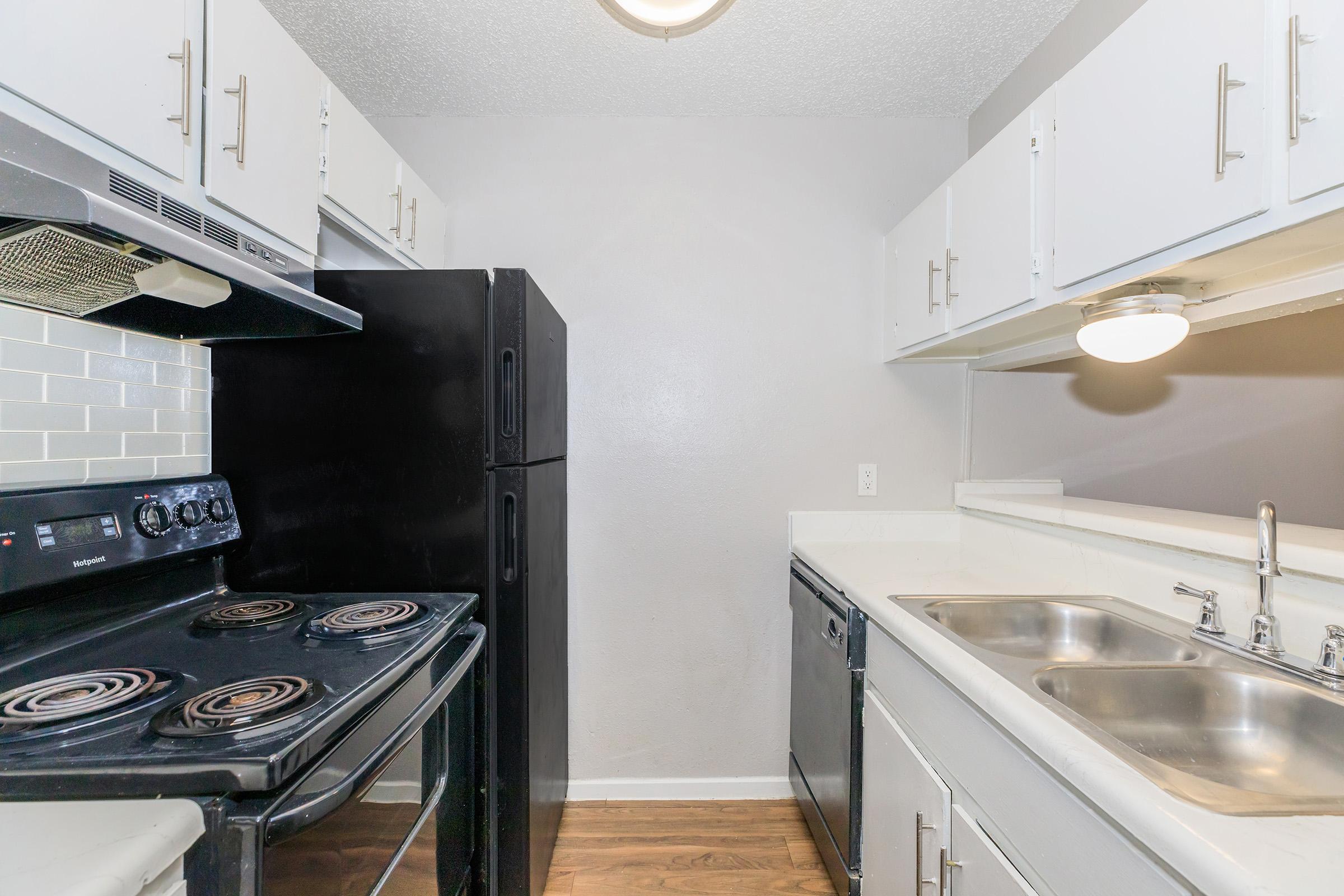 A small kitchen featuring a black refrigerator and a black stove with an oven. There’s a white countertop with a double sink and a dishwasher below white cabinets. The walls are painted a light gray, and the floor has a wood-like finish. Ceiling light is visible, providing bright illumination.