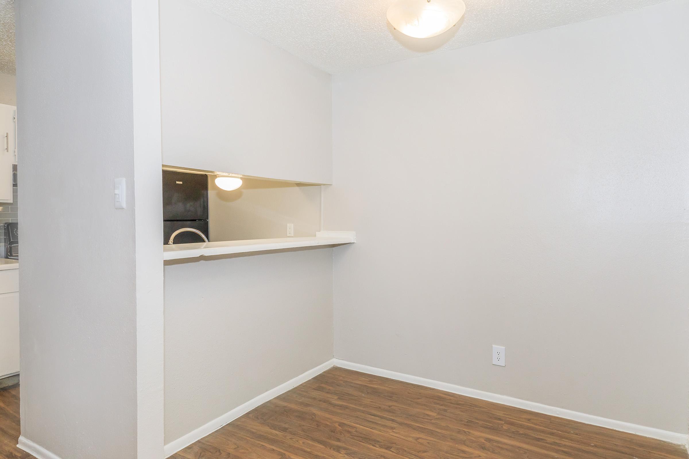 An empty room featuring light gray walls and hardwood flooring. There’s a small opening that leads to a kitchen area, and a ceiling light is installed. The space is uncluttered and ready for furnishings.