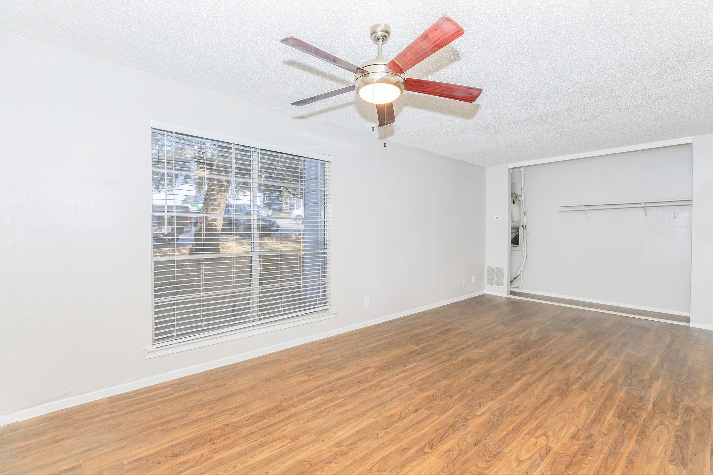 Spacious living room featuring light-colored walls, a large window with blinds, a ceiling fan with wooden blades, and hardwood-style flooring. A corner shelving space is visible, providing additional storage options. The overall ambiance is bright and welcoming.