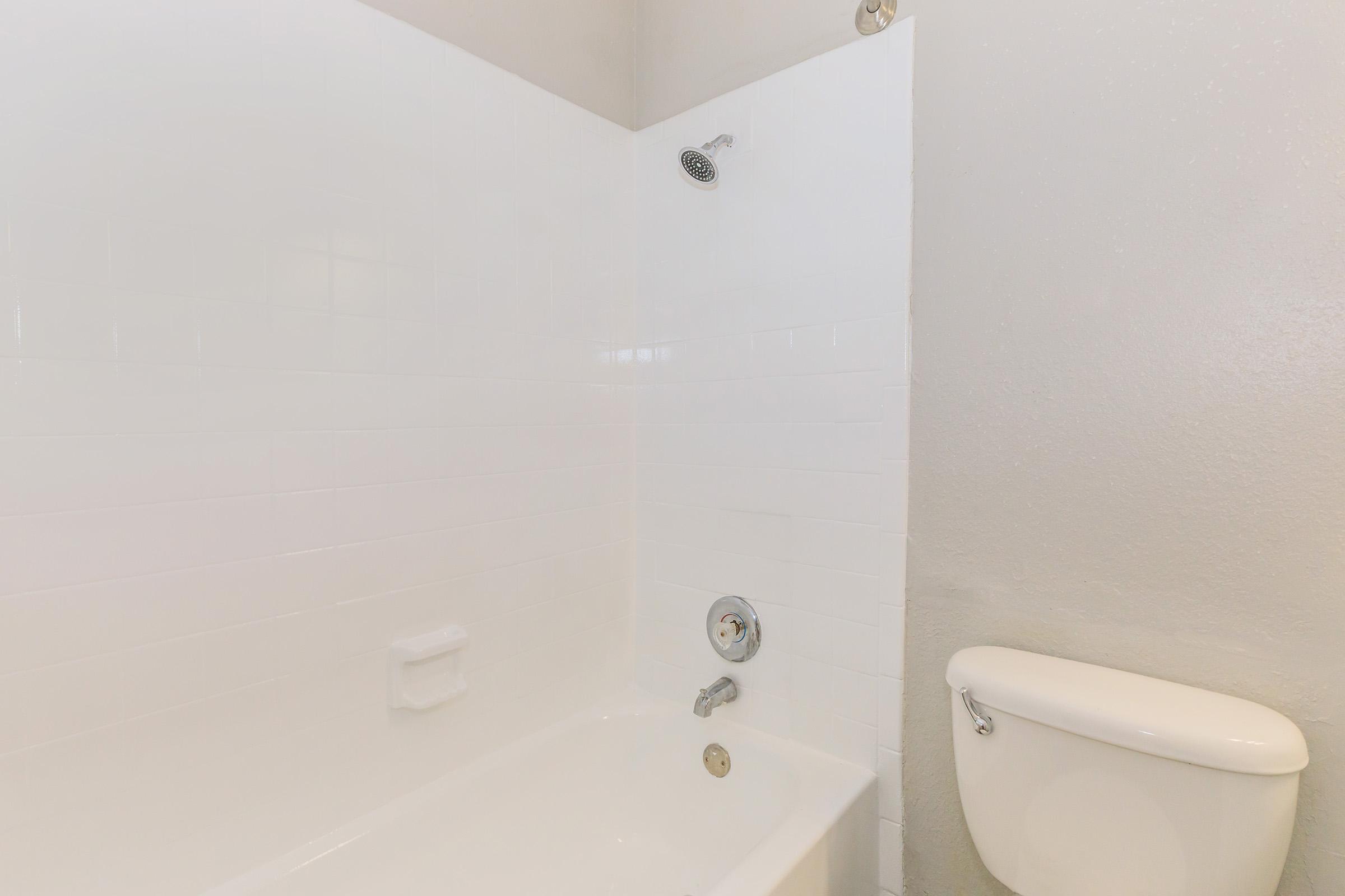 A clean bathroom featuring a white tiled shower area with a showerhead, a bathtub with a faucet, and a white toilet nearby. The walls are light-colored, contributing to a bright and minimalist aesthetic.