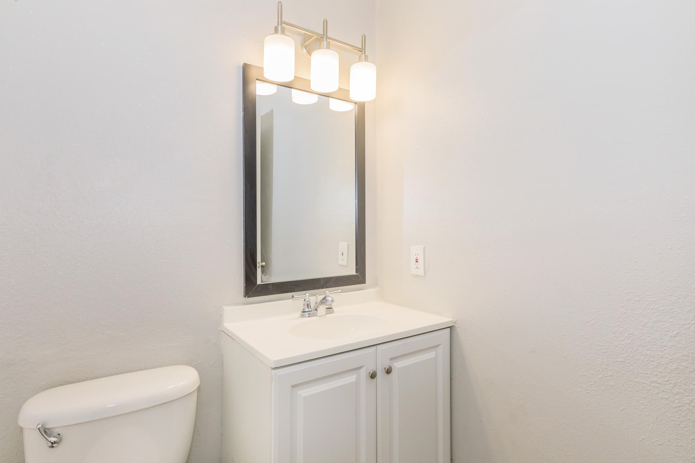A clean, modern bathroom featuring a white vanity with a sink and cabinets, a rectangular mirror above it, and three light fixtures. The wall is painted in a light color, and a toilet is visible in the corner. The overall aesthetic is simple and contemporary.