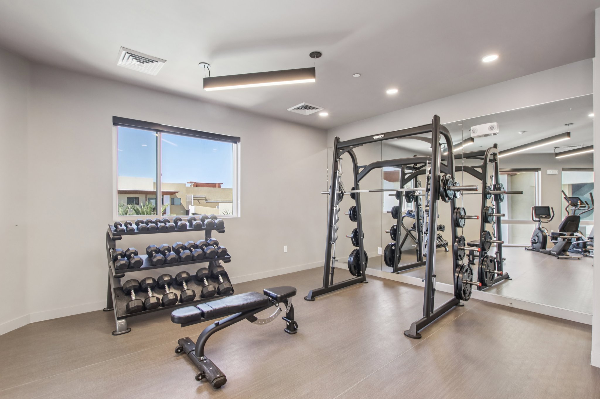 A modern fitness room featuring a rack of dumbbells, a weight bench, and a multi-use strength training machine. Large windows allow natural light to fill the space, and mirrors create a sense of openness. Overall, the design promotes a welcoming and functional workout environment.
