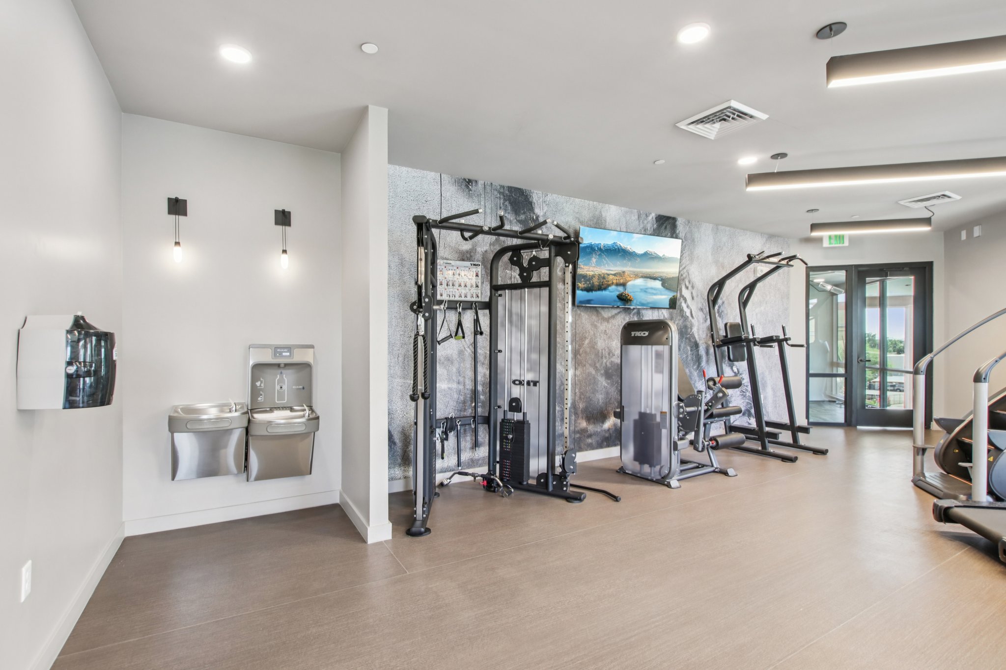 A modern gym interior featuring various exercise equipment, including weight machines and a stationary bike. There are two water fountains on the left, and a large wall-mounted screen displaying a scenic landscape. The floor is light-colored, and natural light enters from a glass door in the background.