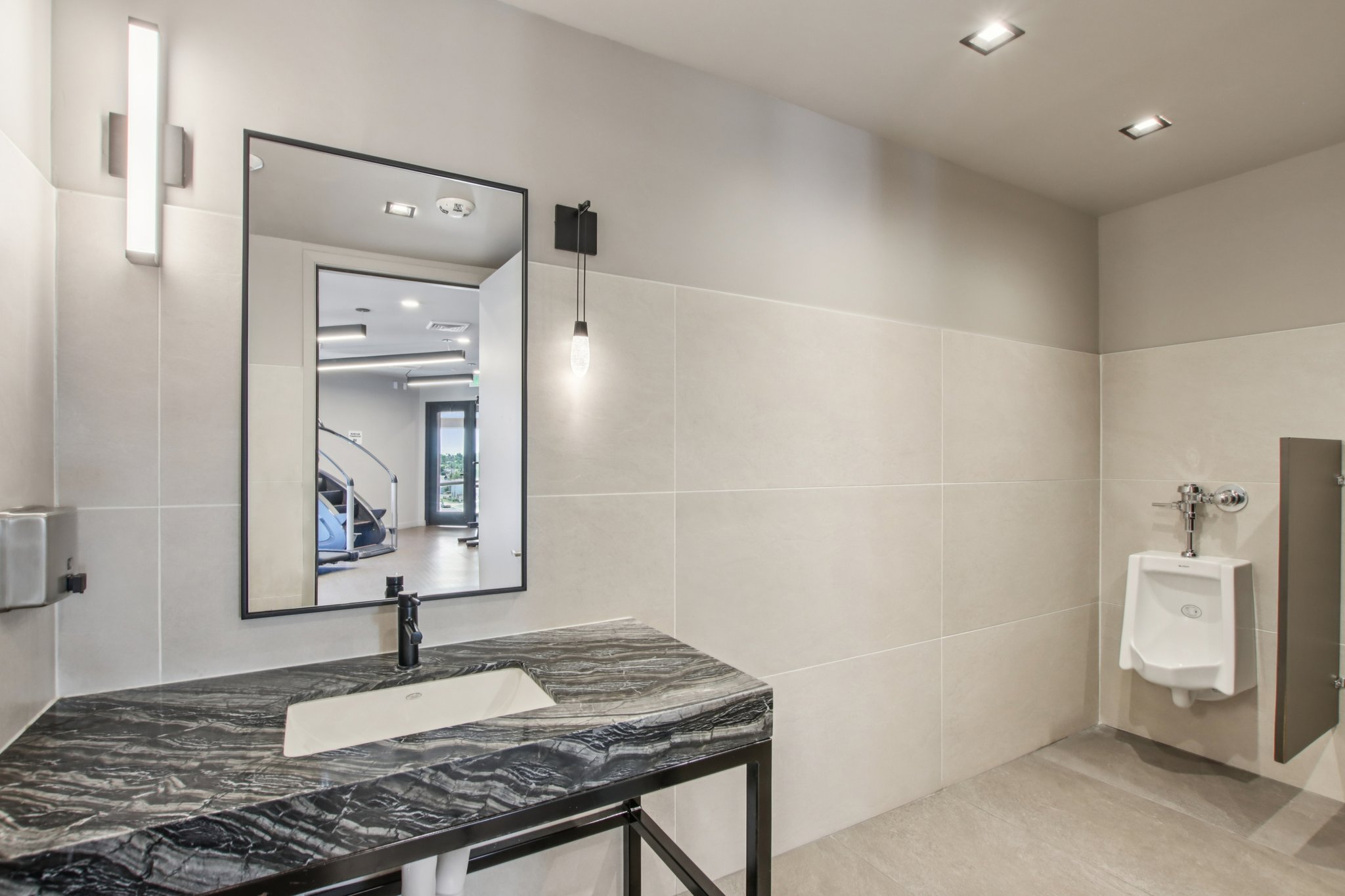 A modern bathroom featuring a dark stone sink on a black metal frame, a large illuminated mirror, and minimalist lighting. The walls are covered in light-colored tiles, and a urinal is visible in the corner. The space has a contemporary feel with clean lines and an open design.