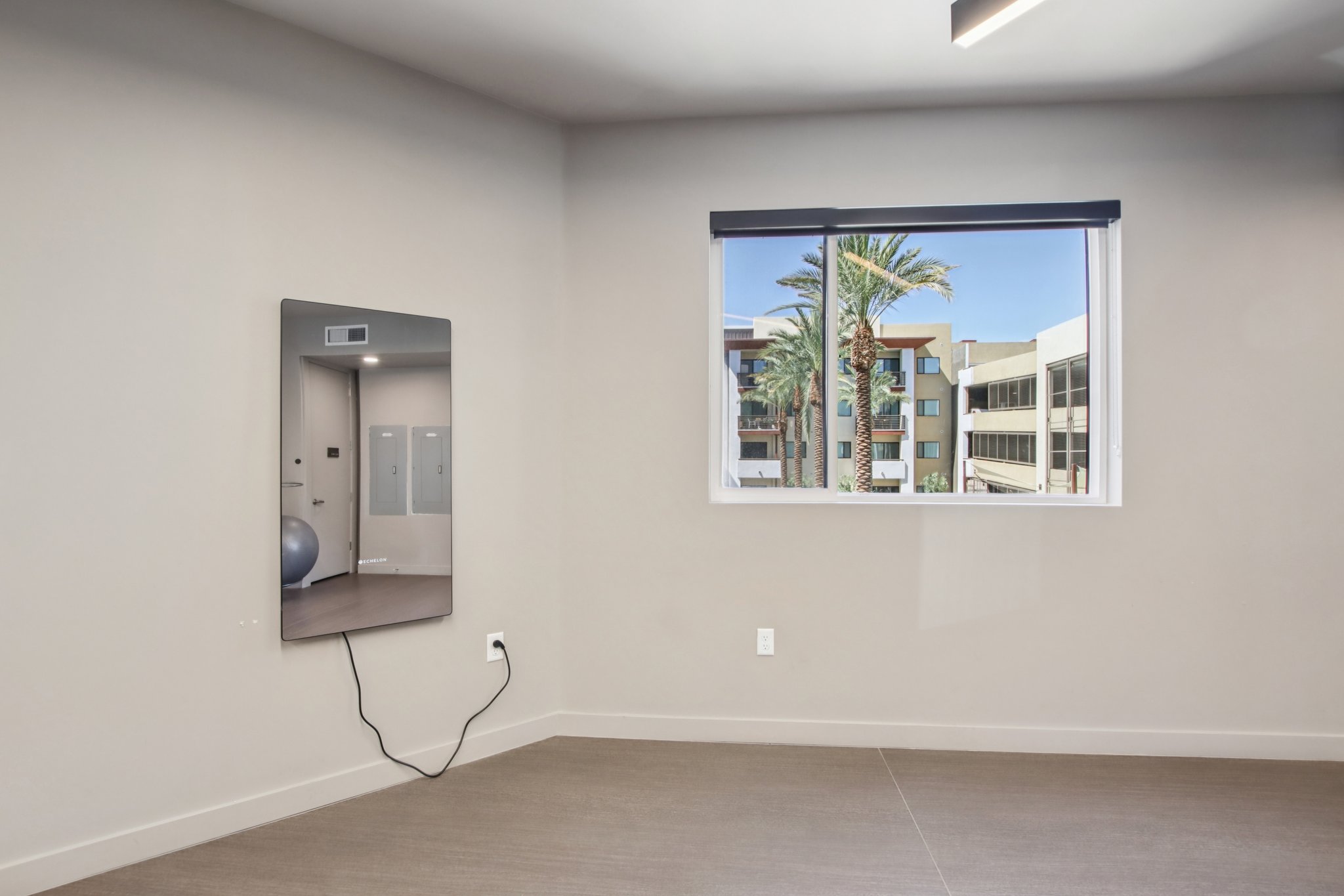 A modern interior space featuring a large mirror mounted on a beige wall, with a view of palm trees and a building visible through a window. The floor is light-colored, and there is a blue exercise ball on the floor, creating a clean and minimalistic atmosphere.