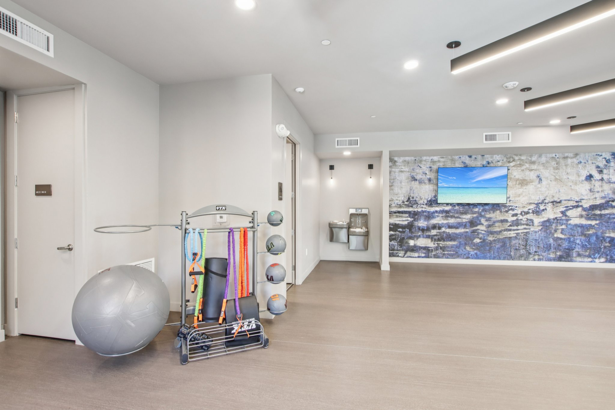 Interior view of a modern fitness studio featuring a gray exercise ball, resistance bands, and a wall-mounted TV displaying a scenic beach image. The walls are a light color with a textured accent wall, and there is a water fountain nearby, contributing to an inviting workout environment.