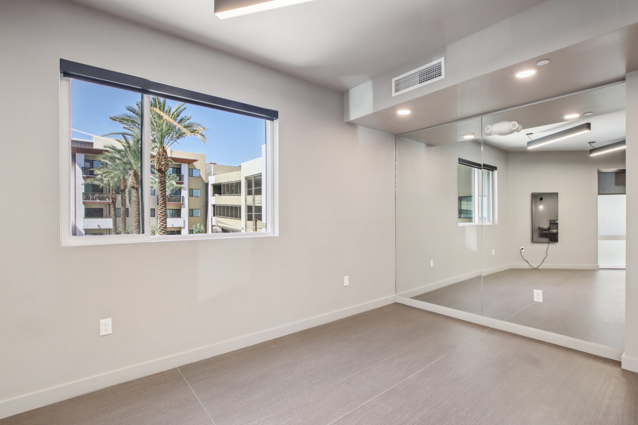 Interior of a modern room featuring a large window with a view of palm trees and a building exterior. One wall is mirrored, reflecting the room's light and spaciousness. The floor is light-colored tile, and there are recessed lights in the ceiling, creating a bright atmosphere.