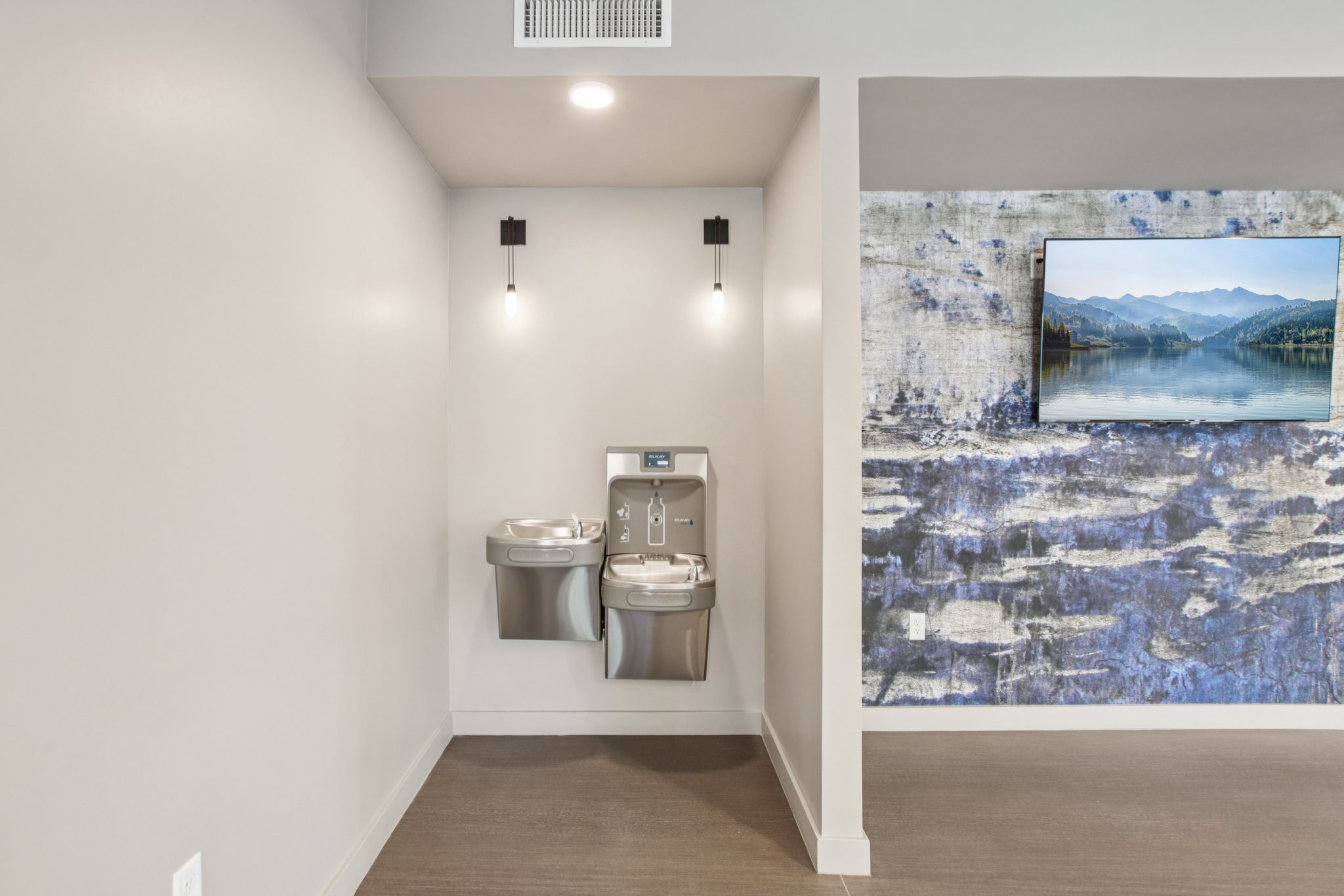 Two modern water fountains are mounted on a light gray wall, illuminated by sconces on either side. To the right, a large flat-screen TV is mounted on a textured blue and gray wall, featuring a serene landscape of mountains and water. The flooring is simple and understated.