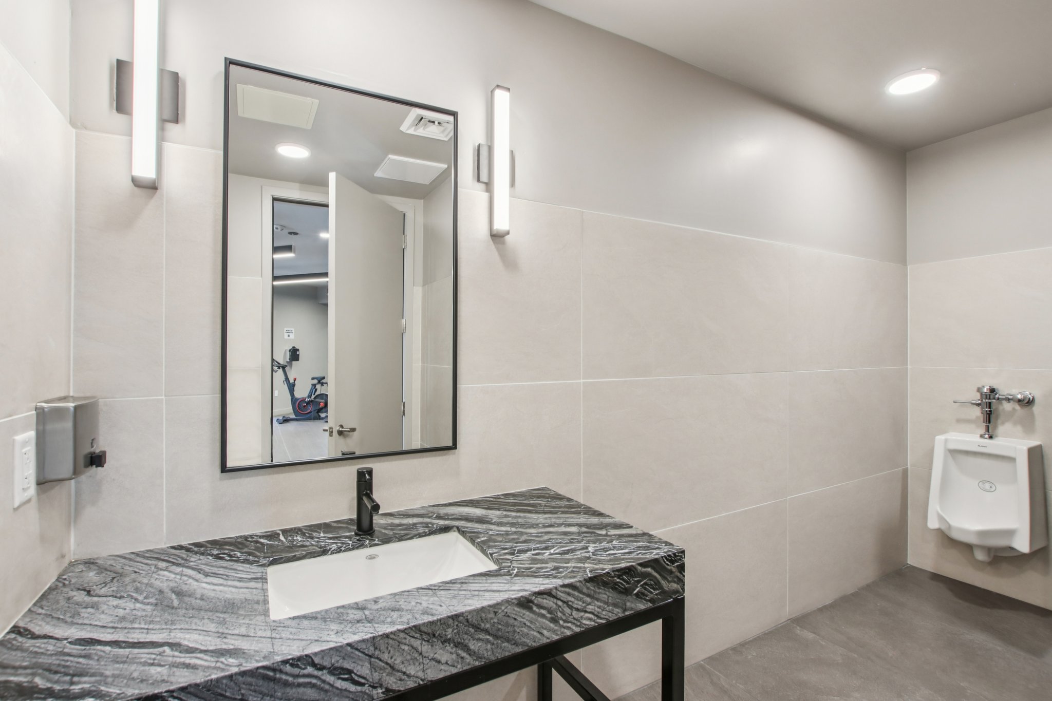 Modern restroom interior featuring a sleek black marble countertop with a rectangular sink, a wall-mounted faucet, and a large mirror. Soft lighting from wall sconces creates a bright ambiance. A urinal is visible on the right side, and a door leading to another area is partially open in the background.