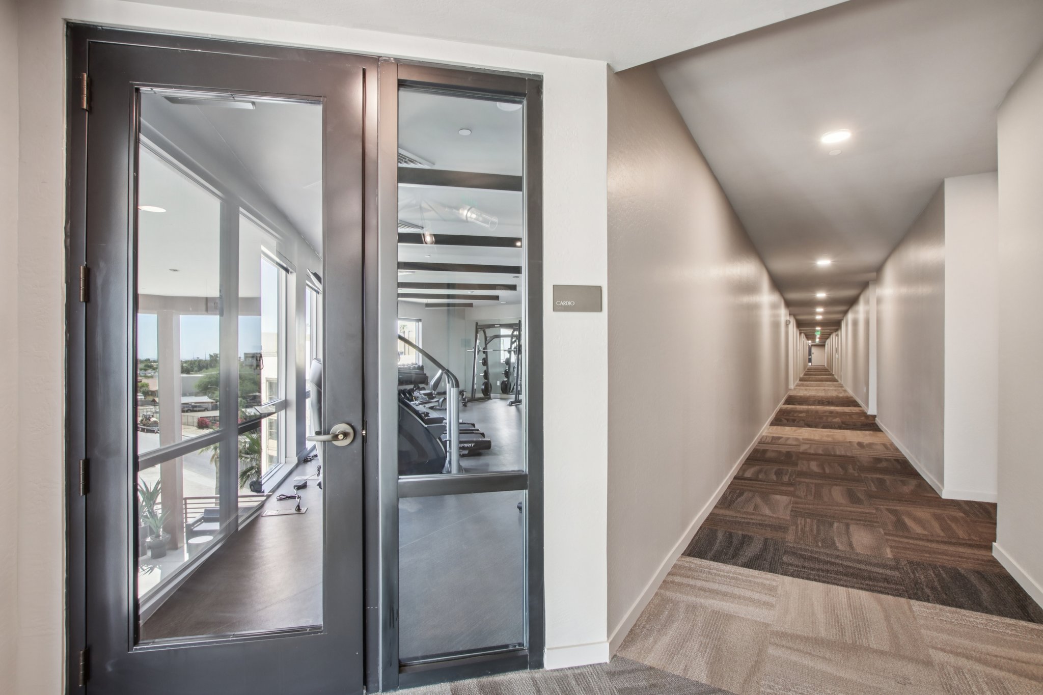A hallway leading to a glass door that opens into a well-lit room with fitness equipment. The hallway features neutral-colored walls and patterned carpet. Natural light streams in through windows on the left side, creating a bright atmosphere.