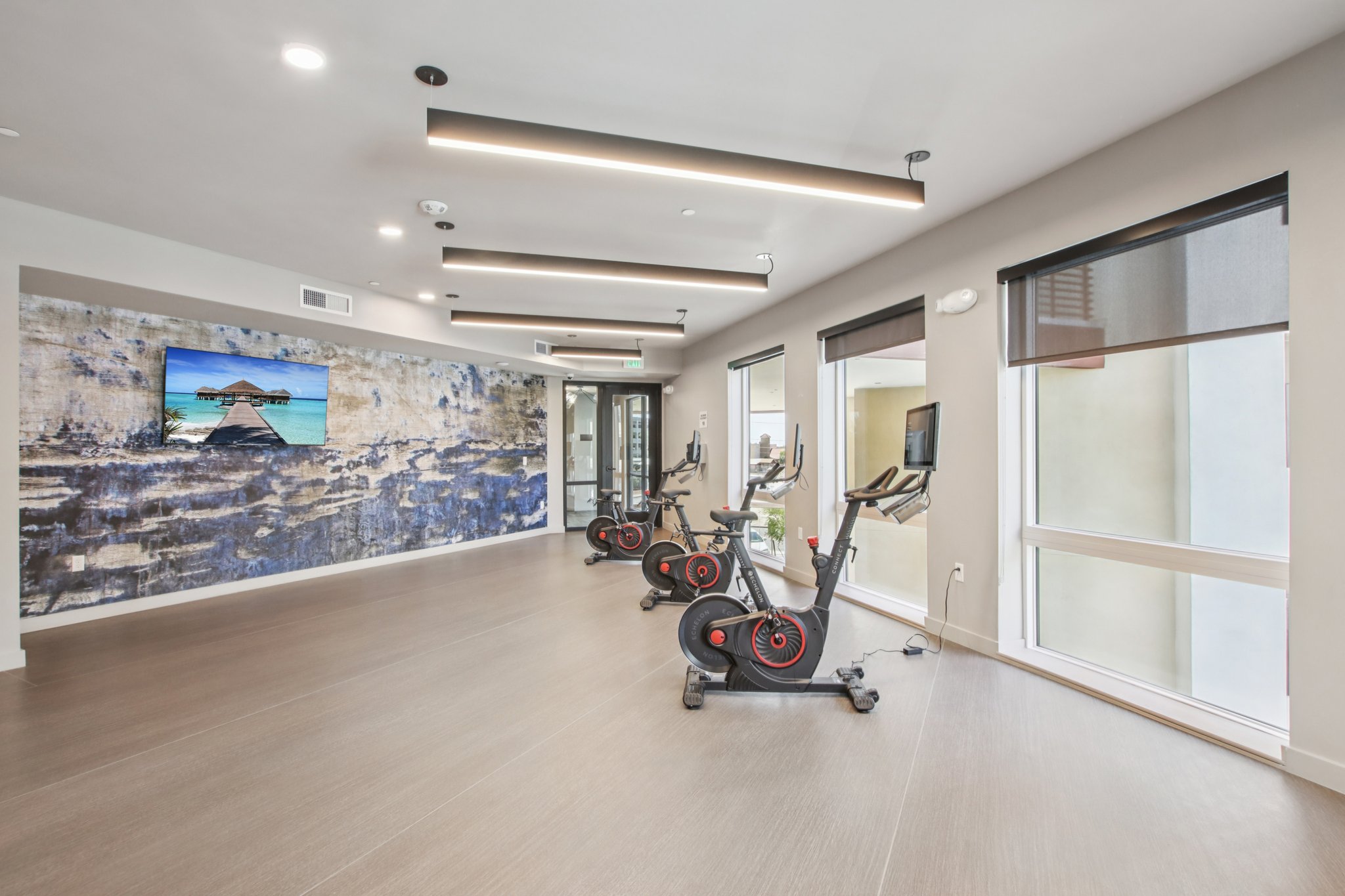 A modern gym interior featuring three stationary bikes in front of a wall with a large mural. The space is bright with natural light, large windows, and contemporary lighting fixtures overhead. A TV mounted on the wall displays a serene landscape image, adding to the inviting atmosphere.