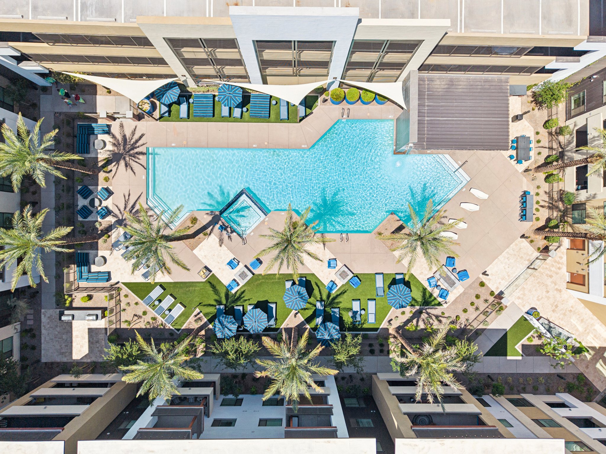 Aerial view of a modern apartment complex featuring a large swimming pool surrounded by palm trees, lounge chairs, and shaded seating areas. The pool area is designed with a unique shape, bordered by manicured lawns and pathways. The surrounding architecture is contemporary, with a mix of greenery and outdoor spaces.