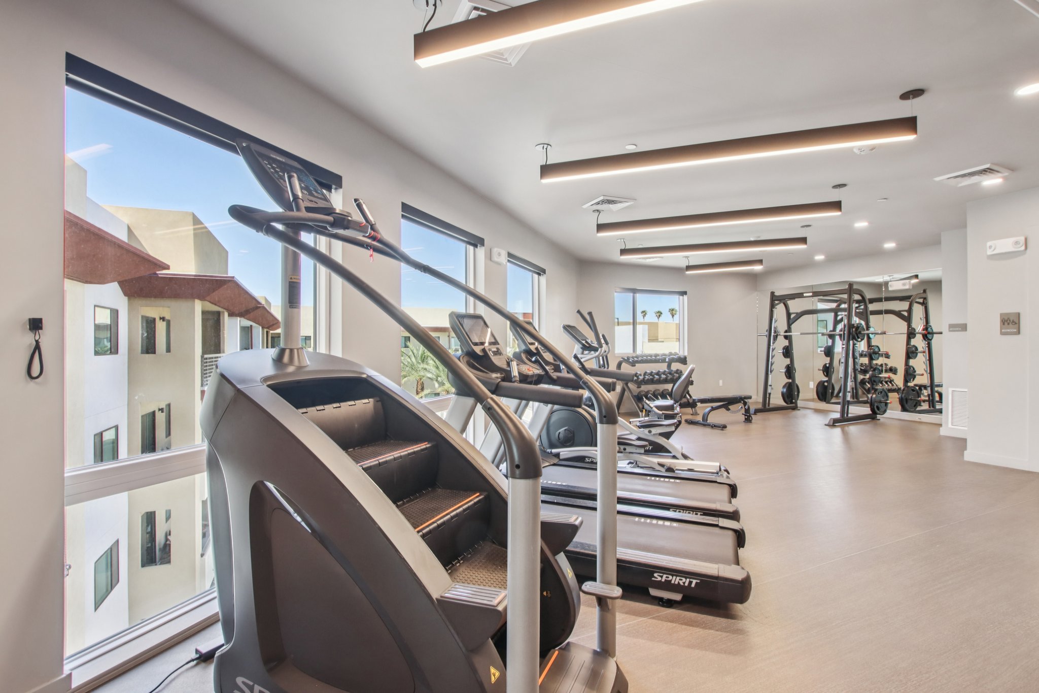 A modern gym interior featuring several cardio machines, including treadmills and an elliptical machine, alongside a section with free weights and weightlifting equipment. Bright, natural light enters through large windows, providing a view of the outside. The space is clean and well-organized.