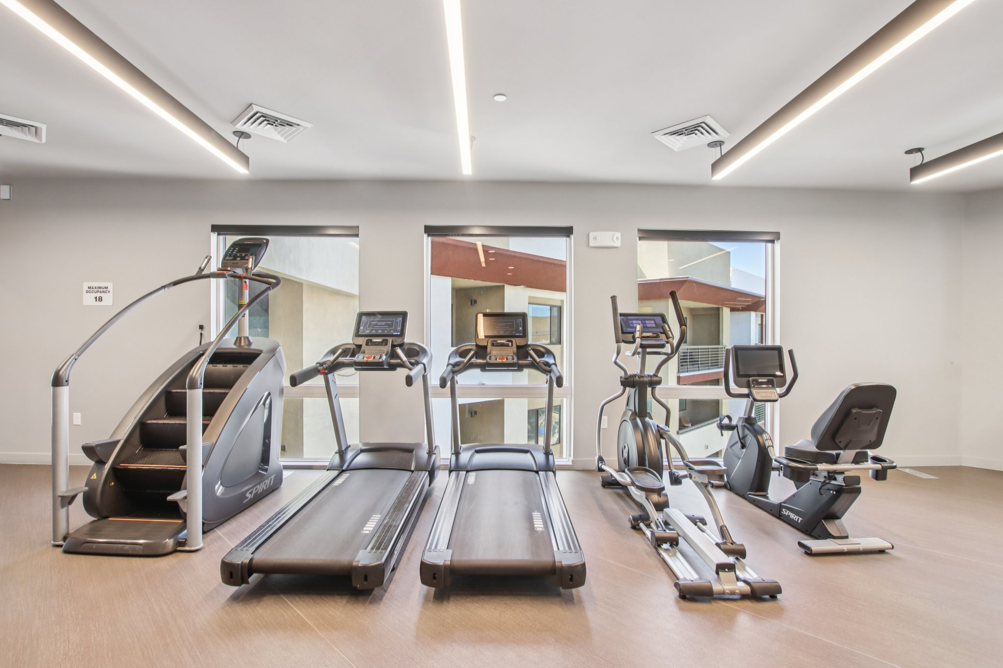 A modern gym featuring various exercise equipment, including a stair climber, treadmills, an elliptical machine, and a stationary bike, all situated in a well-lit room with large windows and a neutral color scheme.