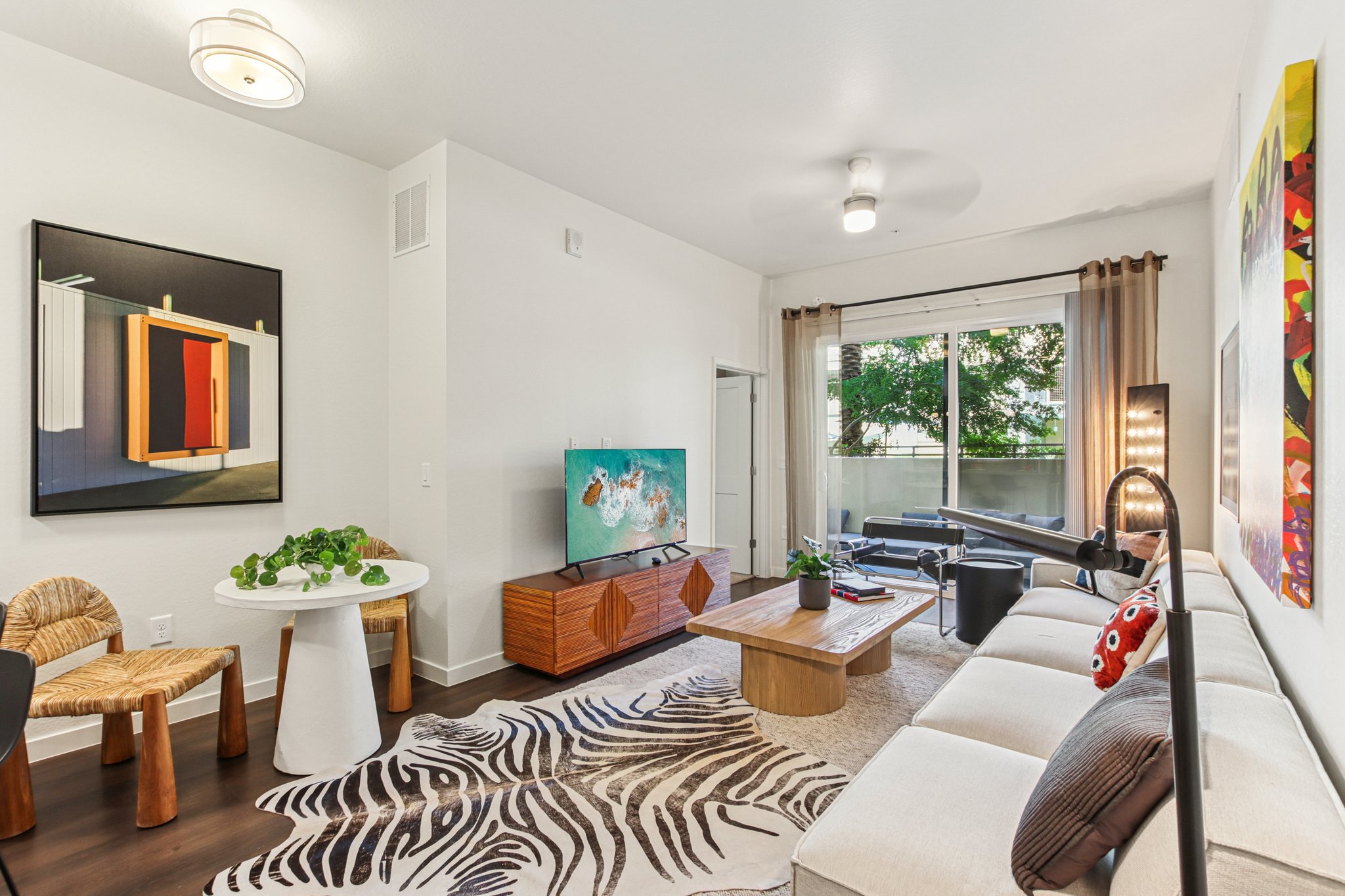 A modern living room featuring a beige couch, a coffee table, and a TV unit. There's a round white dining table with unique chairs, a patterned rug, and artwork on the walls. Large windows allow natural light, and green plants add a touch of nature to the space.