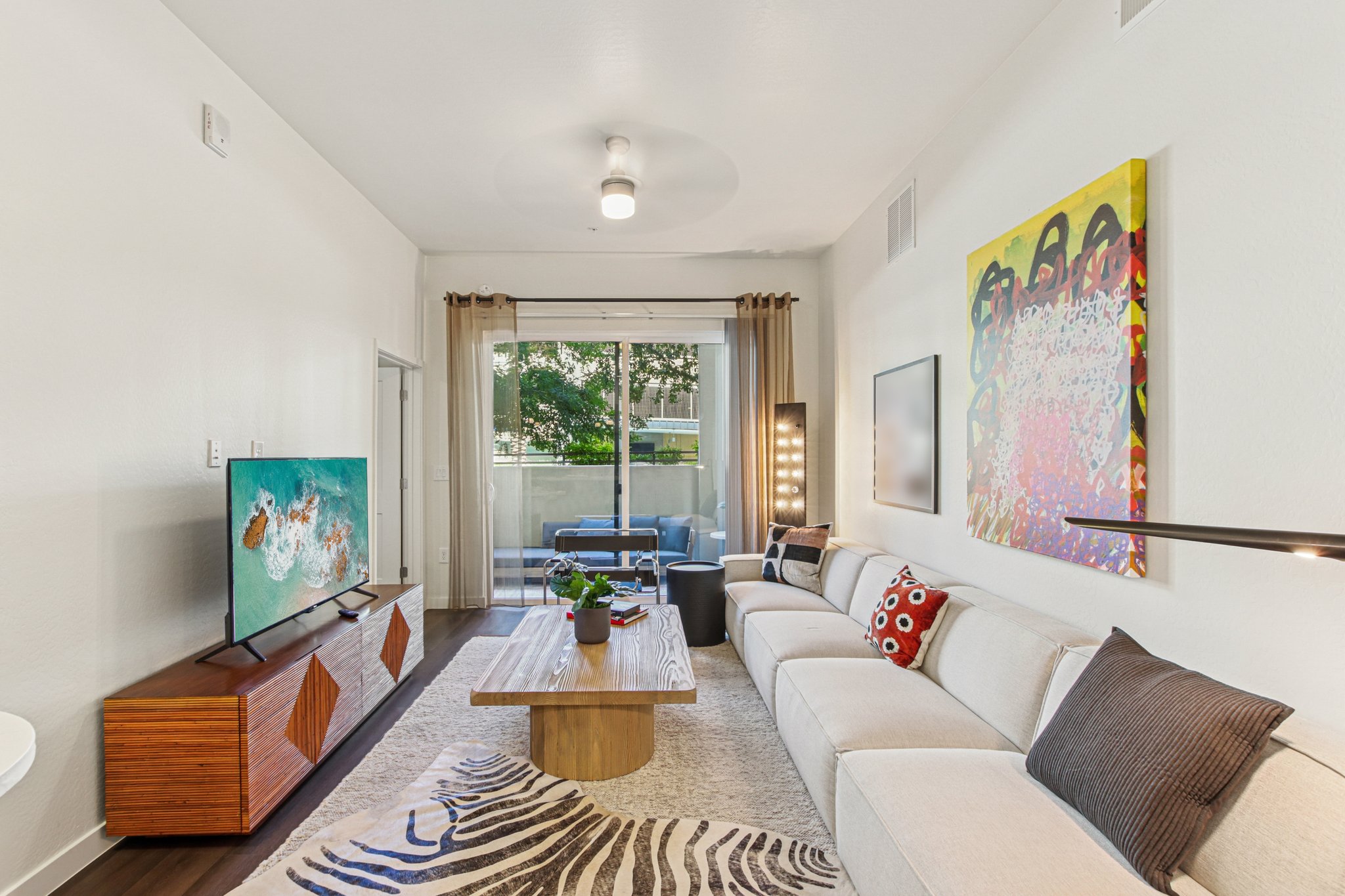 A modern living room featuring a light-colored sofa with patterned cushions, a wooden coffee table on a textured rug, a TV mounted on a stand with artwork beside it, and large windows that let in natural light, showcasing an outdoor space.