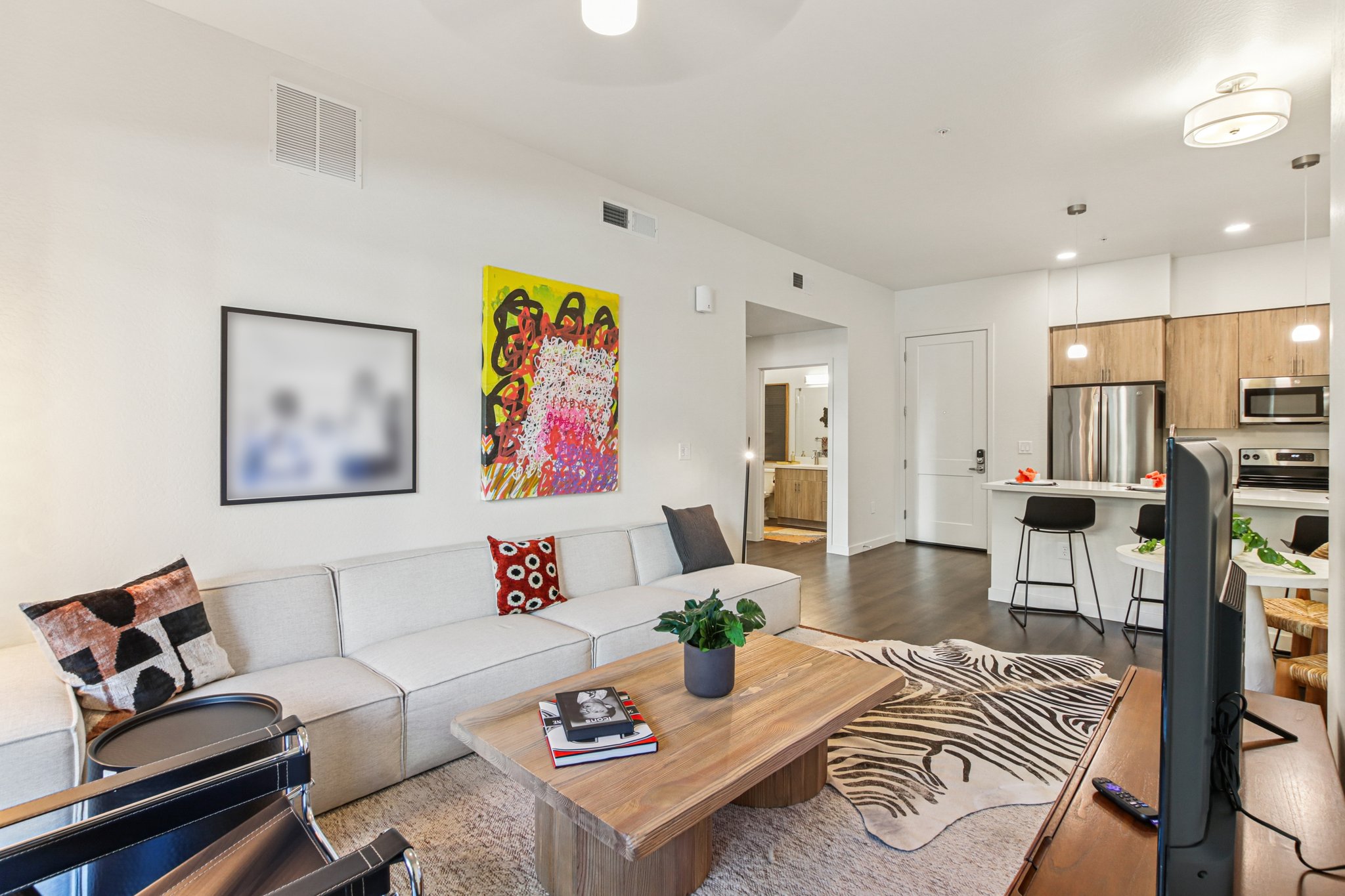 A bright and modern living room featuring a light-colored sectional sofa with decorative pillows, a wooden coffee table on a patterned rug, and a television. A colorful abstract painting hangs on the wall, and the space includes a dining area with bar stools. The kitchen is visible in the background.