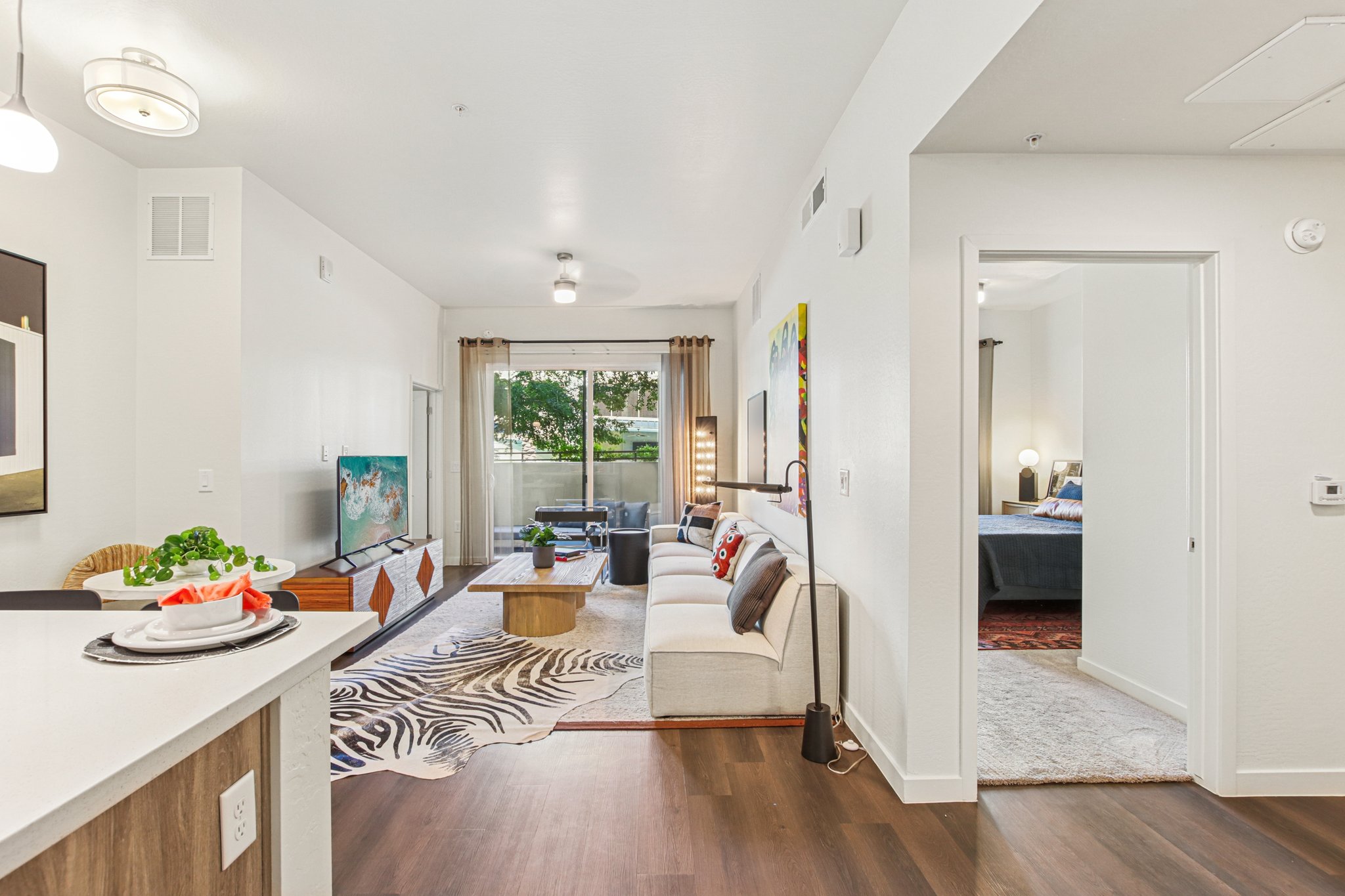 A modern living space featuring a bright and airy layout. The area includes a stylish sofa with decorative pillows, a coffee table, and a television. There are large windows allowing natural light and a view of greenery. The adjacent room leads to a cozy bedroom with tasteful decor.