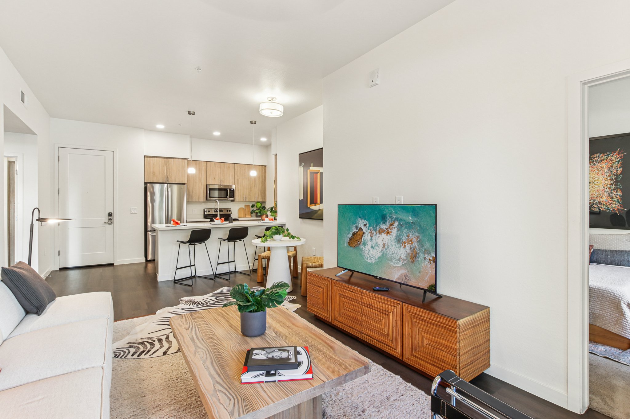 A modern living space featuring a light-colored couch, a wooden coffee table, and a flat-screen TV on a wooden cabinet. The open kitchen in the background includes bar stools and contemporary appliances. Natural light brightens the room, accented by plants and decorative artwork.