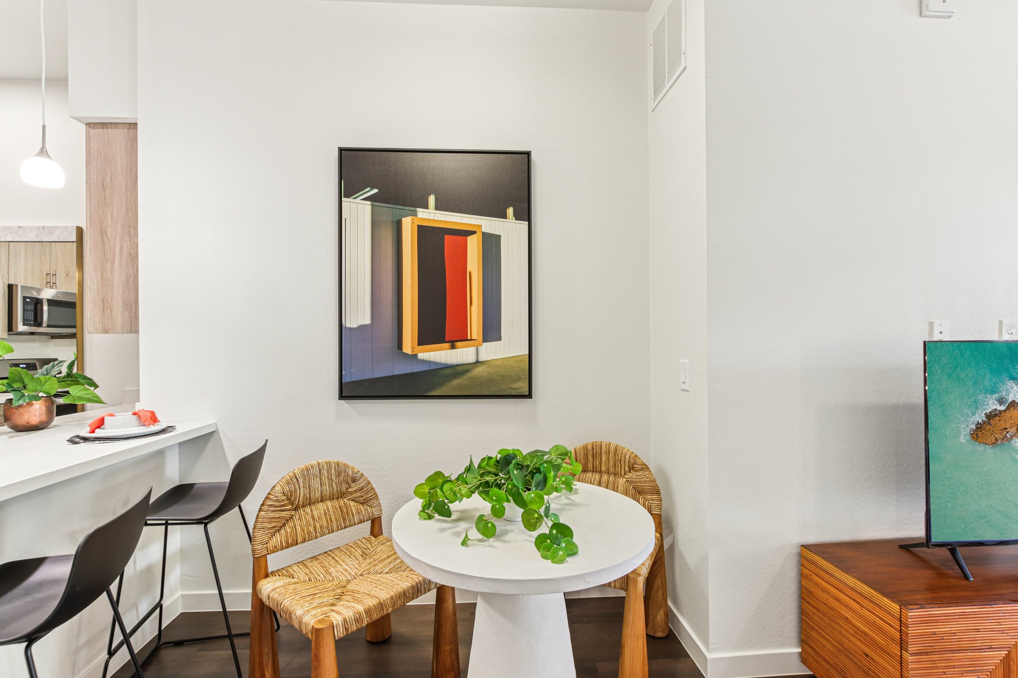 A modern dining space featuring a round white table with a plant centerpiece, surrounded by two woven chairs. In the background, there's a framed abstract artwork with red and black elements, and a kitchen area with bar stools and contemporary decor. A wooden console holds a decorative screen.