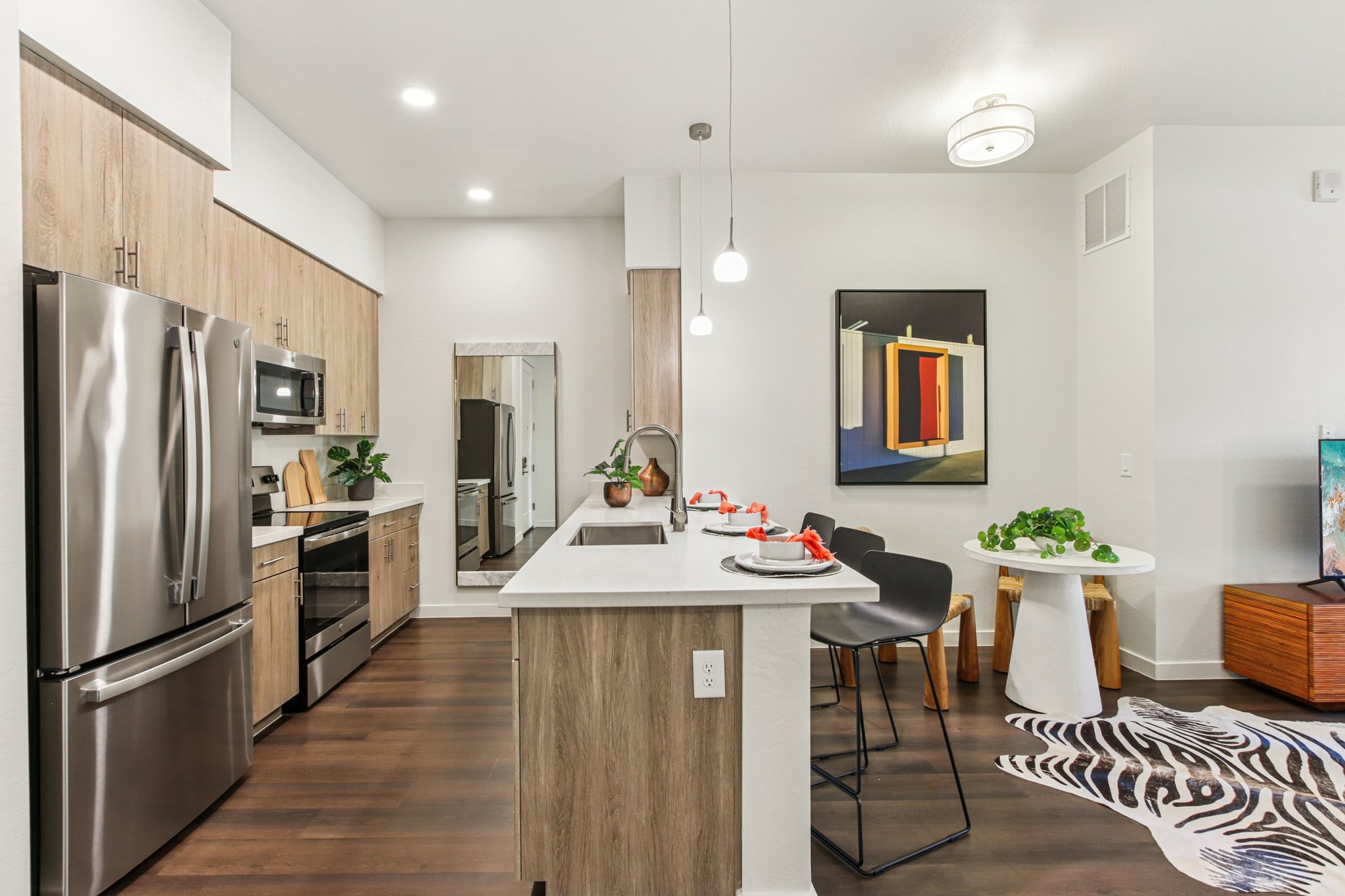 Modern kitchen with stainless steel appliances, including a fridge and microwave. A sleek island features a sink and is surrounded by black bar stools. A dining table with decorative plates sits nearby, complemented by a colorful abstract painting on the wall and a zebra-patterned rug on the floor.
