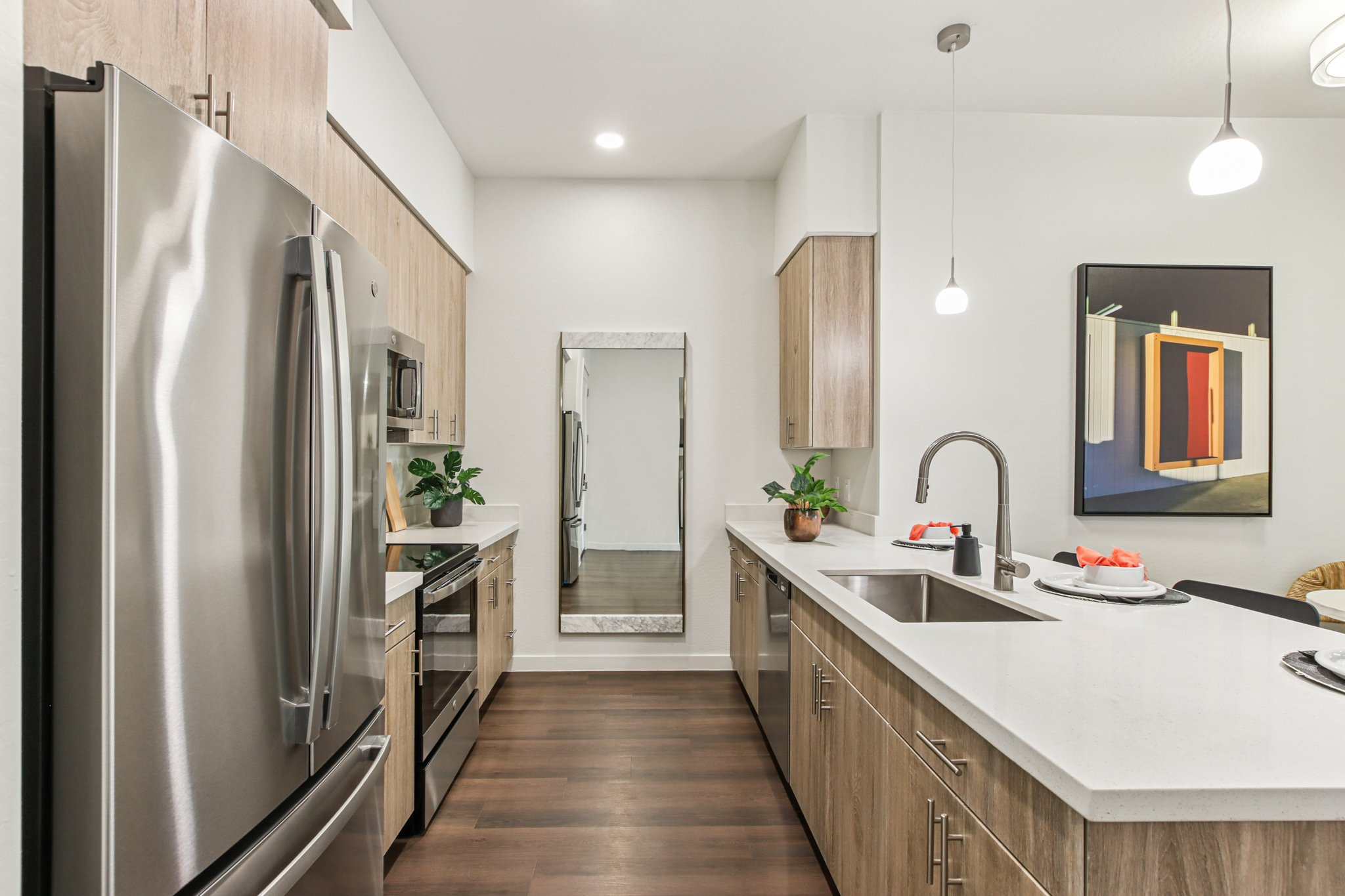 Modern kitchen featuring stainless steel appliances, including a refrigerator and stove. The light wood cabinetry complements the sleek countertops. A central island with a sink is surrounded by a few decorative plants, and minimalistic pendant lights hang above. A mirror reflects the room, adding depth to the space.