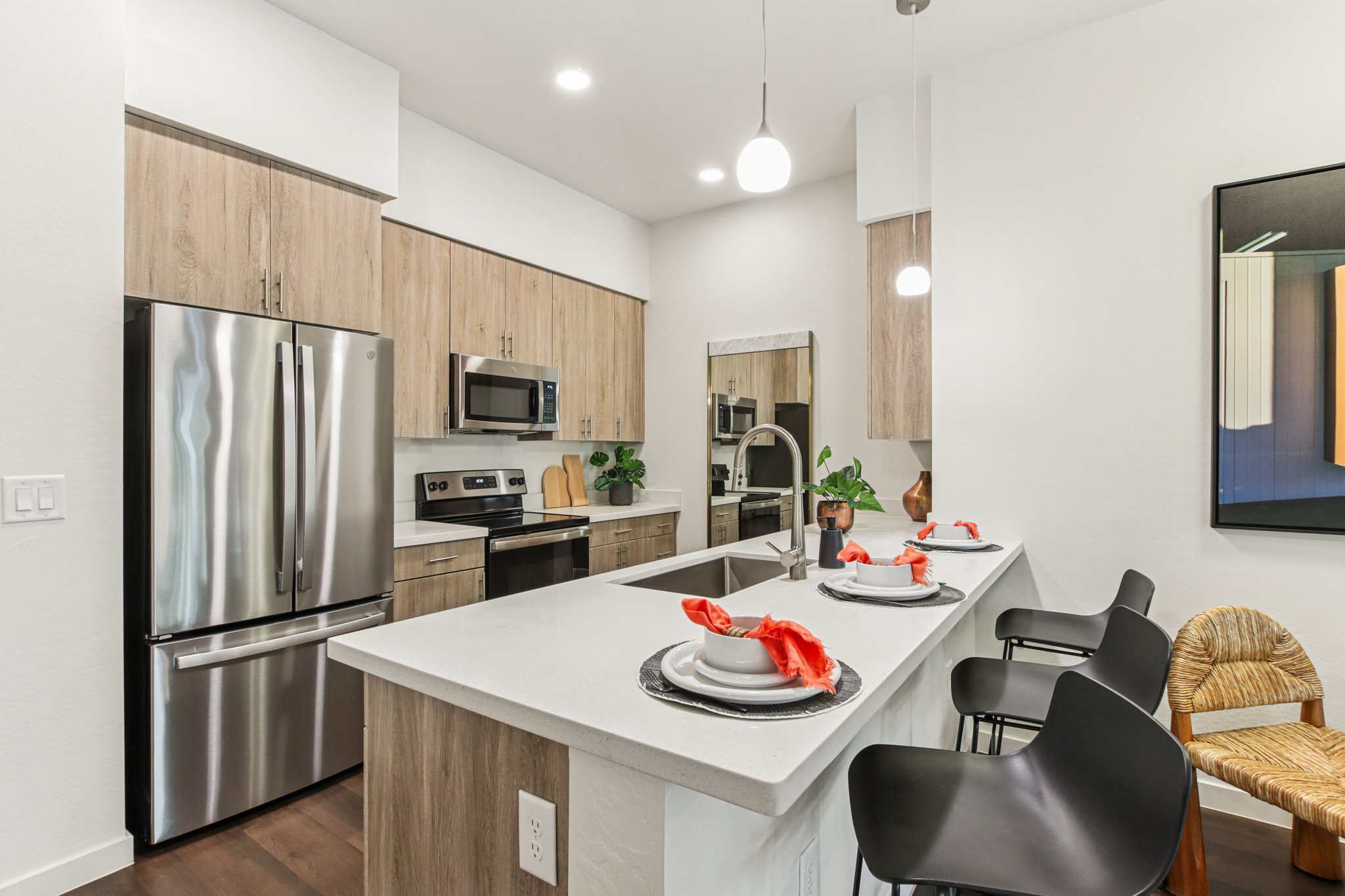 Modern kitchen with light wooden cabinets, stainless steel appliances, a gray countertop, and a central island with bar stools. A neatly set dining table features plates and decorative napkins. The space is well-lit with pendant lights and has stylish decor, including greenery and wall art.