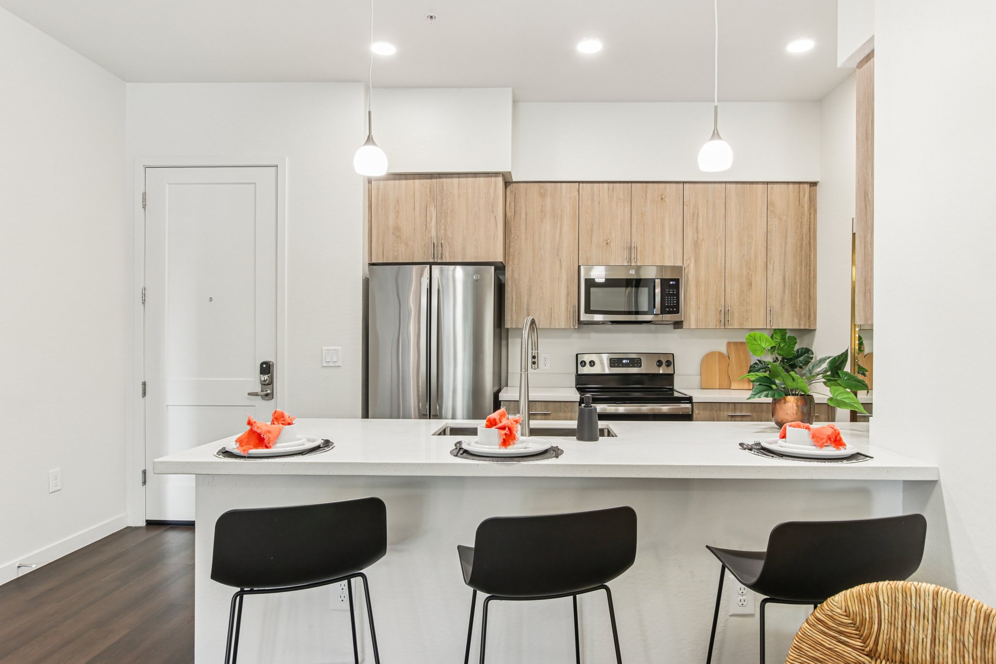 Modern kitchen featuring light wood cabinetry, stainless steel appliances, and a white countertop. Two black barstools are positioned at the island, which is set with decorative plates and vibrant orange accents. Soft lighting from pendant lights enhances the contemporary feel of the space.