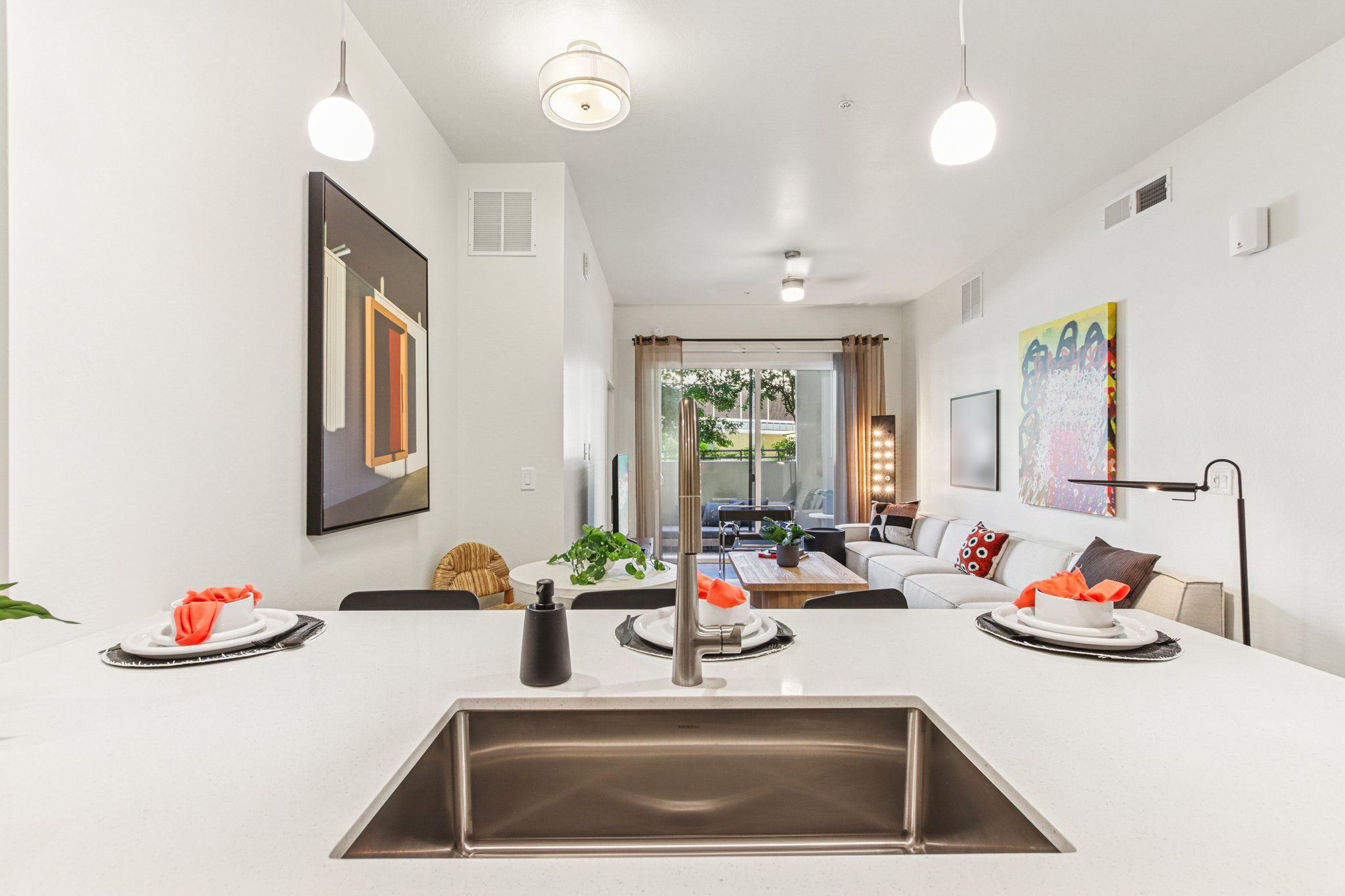 Modern kitchen with a sleek countertop and a sink, featuring neatly arranged plates and utensils. In the background, a cozy living area is visible, furnished with a sofa and decorative lights. Artwork decorates the walls, complementing the bright and airy atmosphere created by large windows.