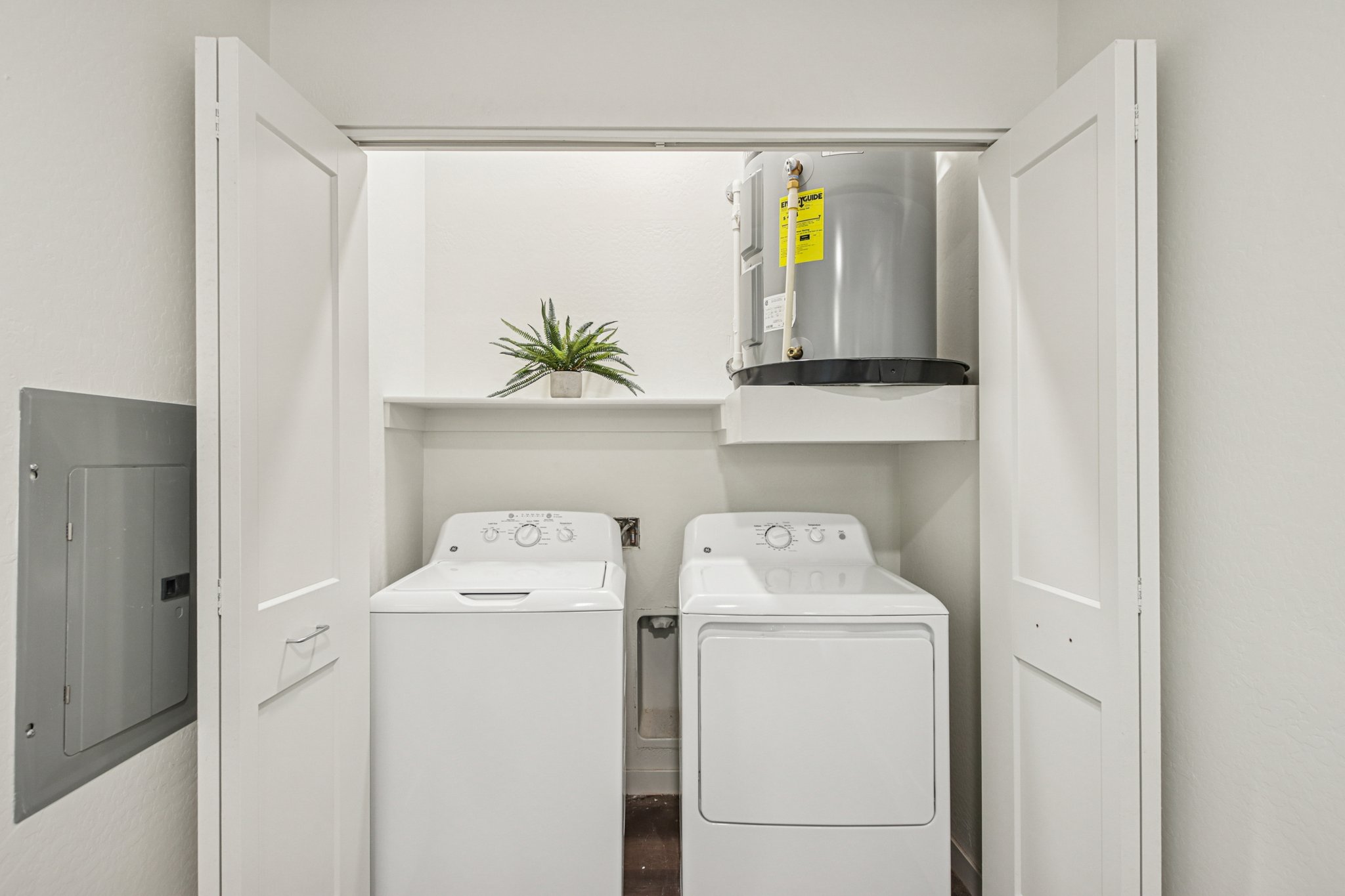 A laundry closet featuring a stacked washer and dryer with a small shelf above. A potted plant is placed on the shelf, and the closet doors are open. A gray water heater is visible above the dryer, and there is a gray electrical panel mounted on the left wall.