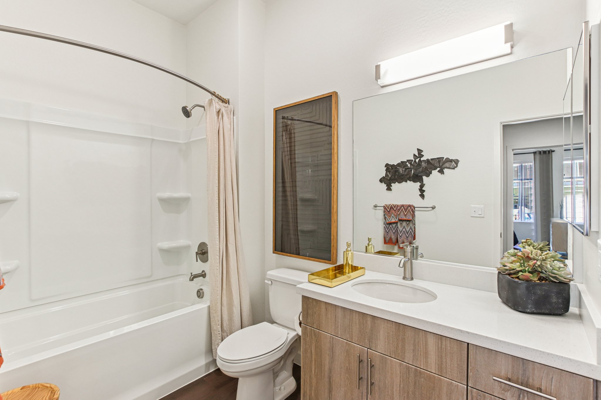 A modern bathroom featuring a white bathtub with a shower, a sleek vanity with a circular sink, and wooden cabinetry. A large mirror reflects the space, and a decorative plant sits on the countertop. The room is well-lit with bright overhead lighting and has a clean, contemporary design.