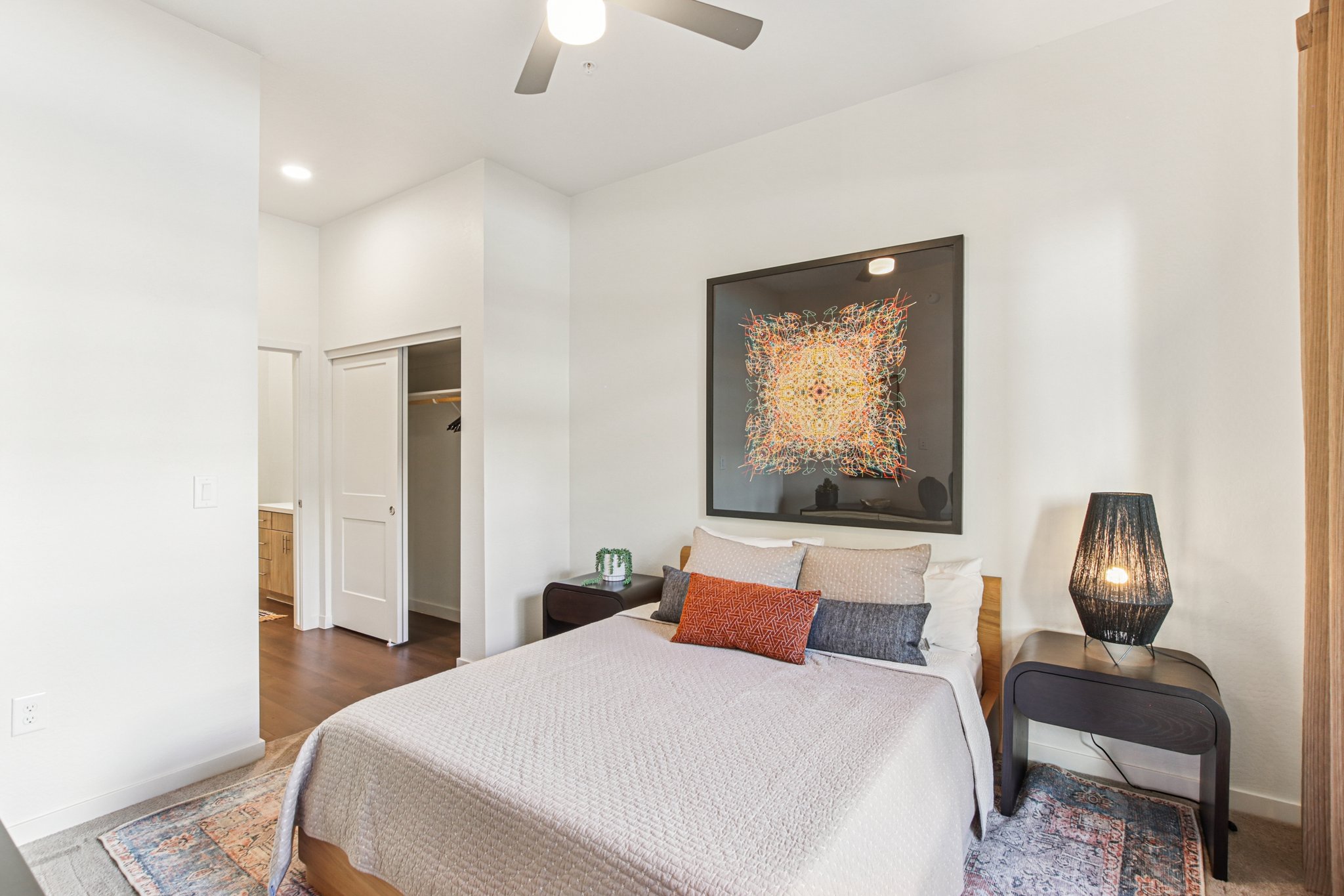 A cozy bedroom featuring a neatly made bed with neutral-colored bedding and an orange accent pillow. A modern artwork hangs above the bed, complemented by a stylish bedside lamp. The room has light-colored walls, a wooden floor, and a closet with a door visible in the background, creating a warm and inviting atmosphere.